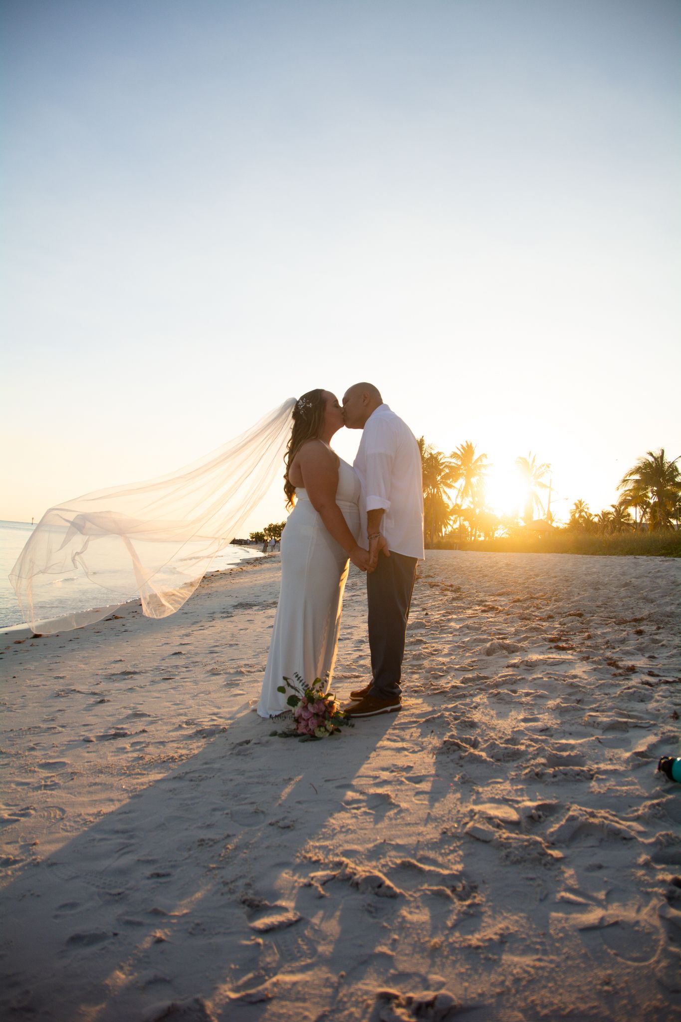 Couple sharing romantic moment at sunset on beach with flowing veil and palm trees in background.