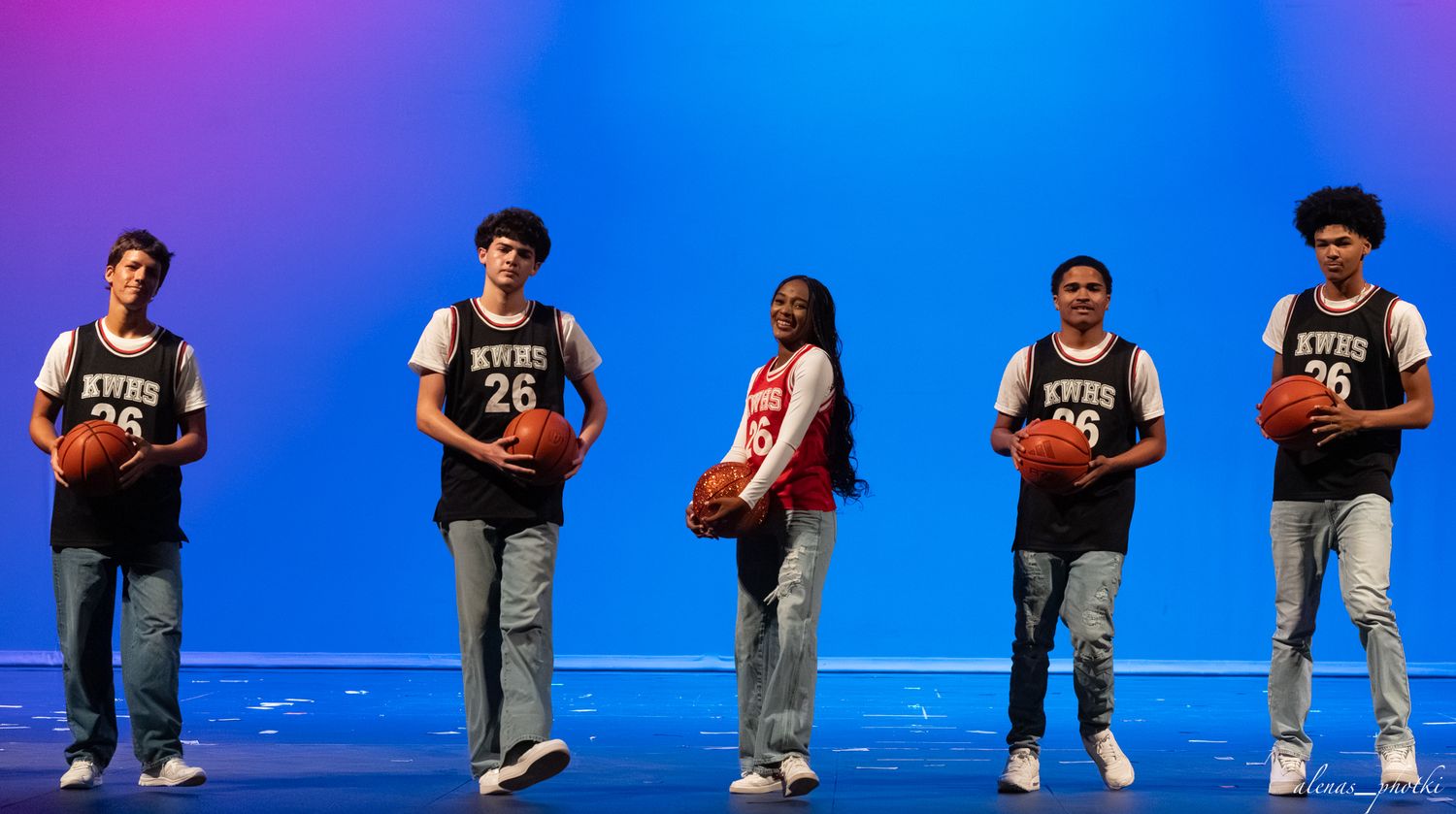 Five teens in matching black basketball jerseys numbered 26 pose on a blue-lit stage holding basketballs.