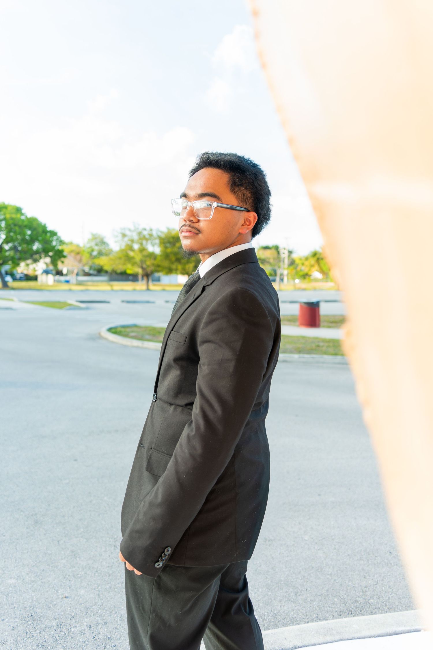 Young Black man in black suit and glasses standing outdoors near a road, looking confident and professional.