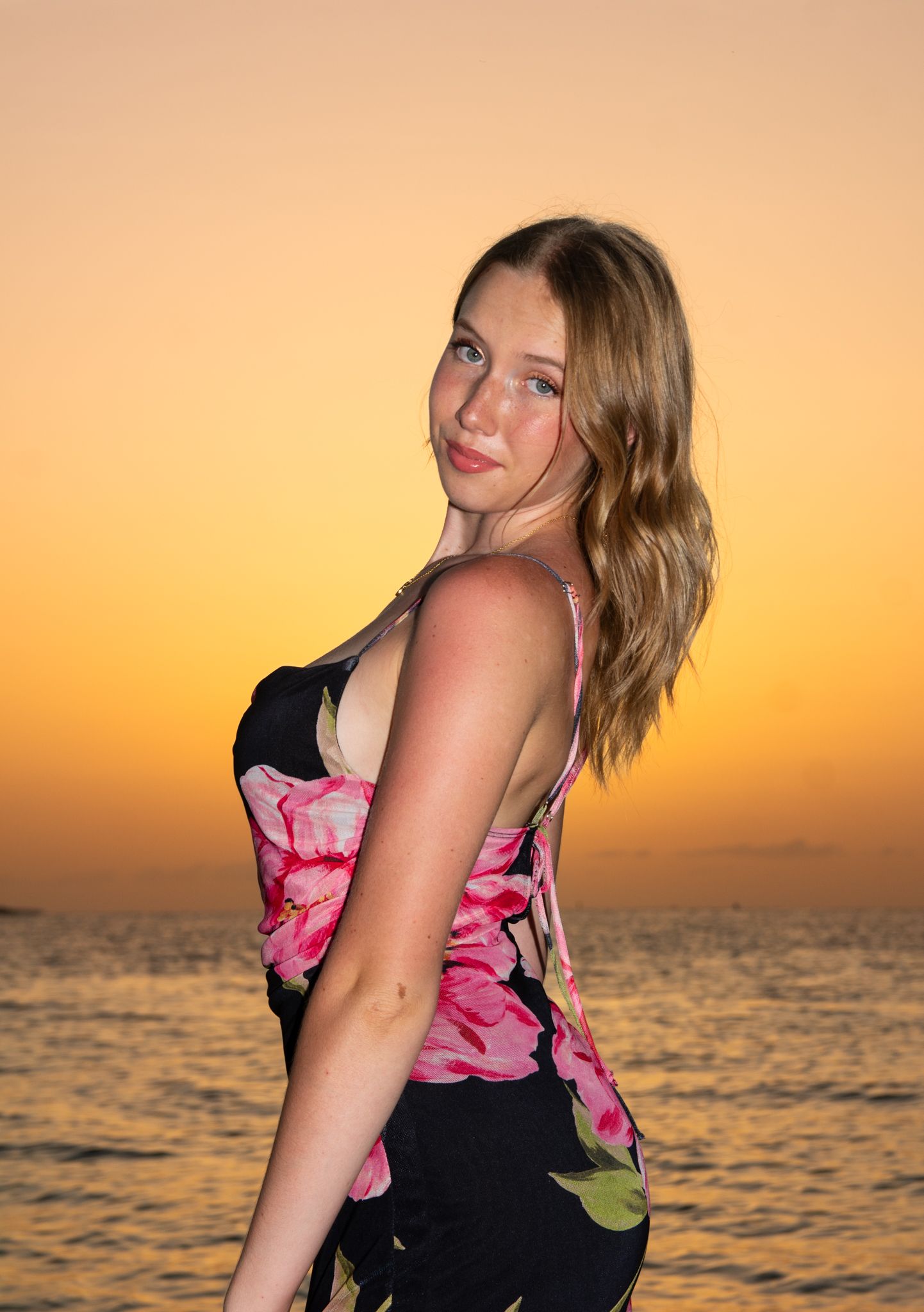 Person in pink and black dress poses by ocean during vibrant sunset.