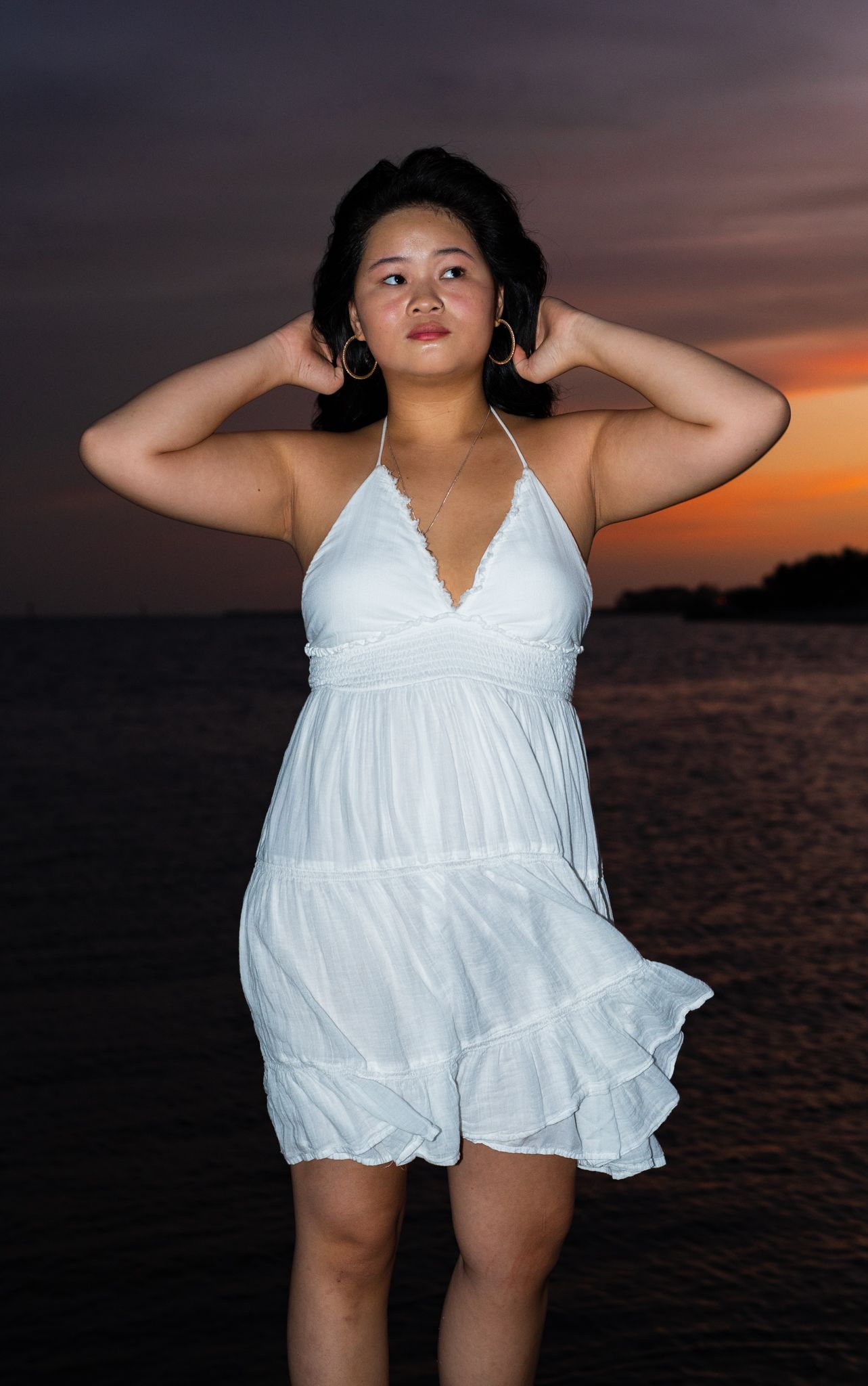 A figure in a flowing white sundress poses against a dramatic sunset over water.
