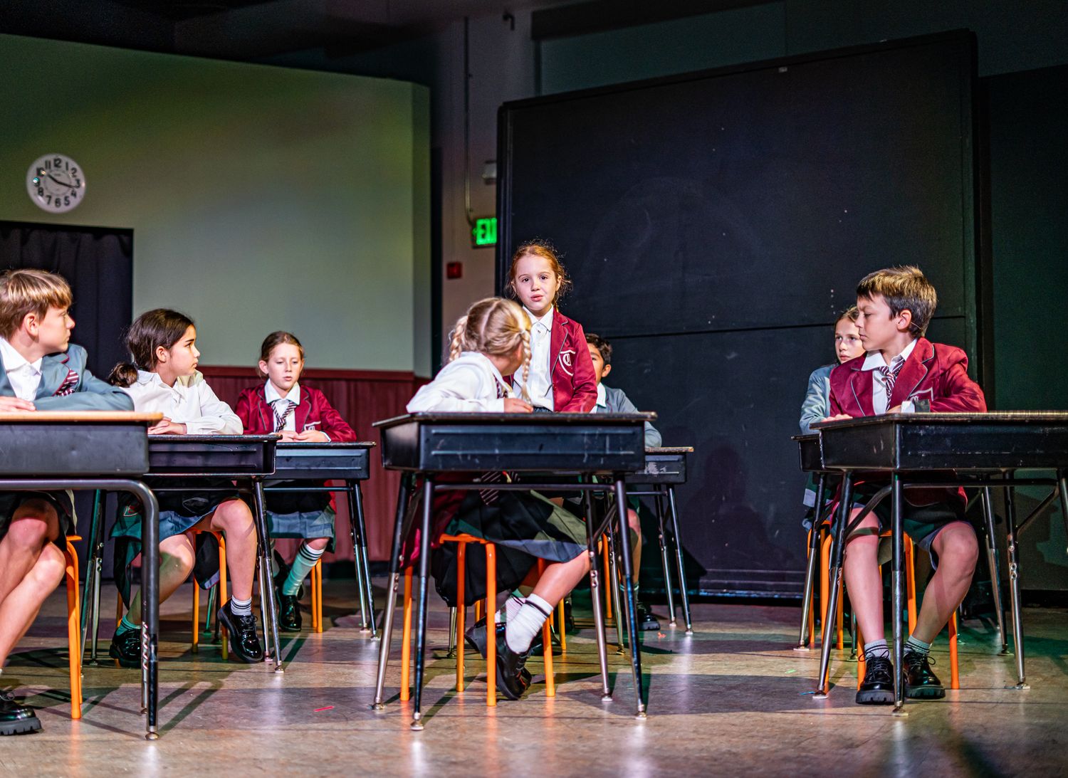 Students in school uniforms sitting at desks during a theatrical classroom scene.