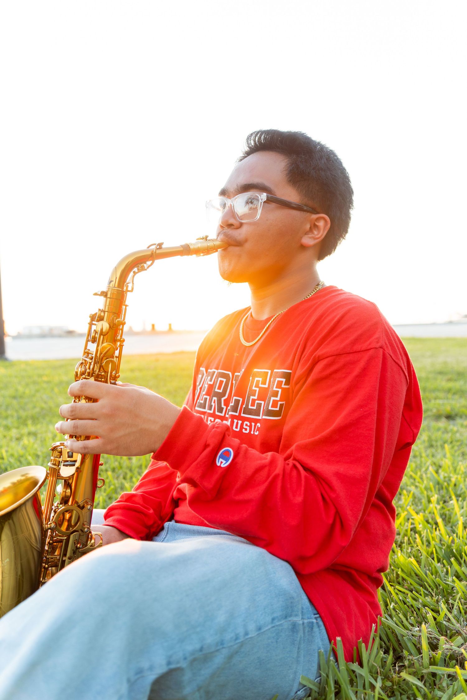 Young man in red sweatshirt playing alto saxophone outdoors on grass during golden hour sunset.