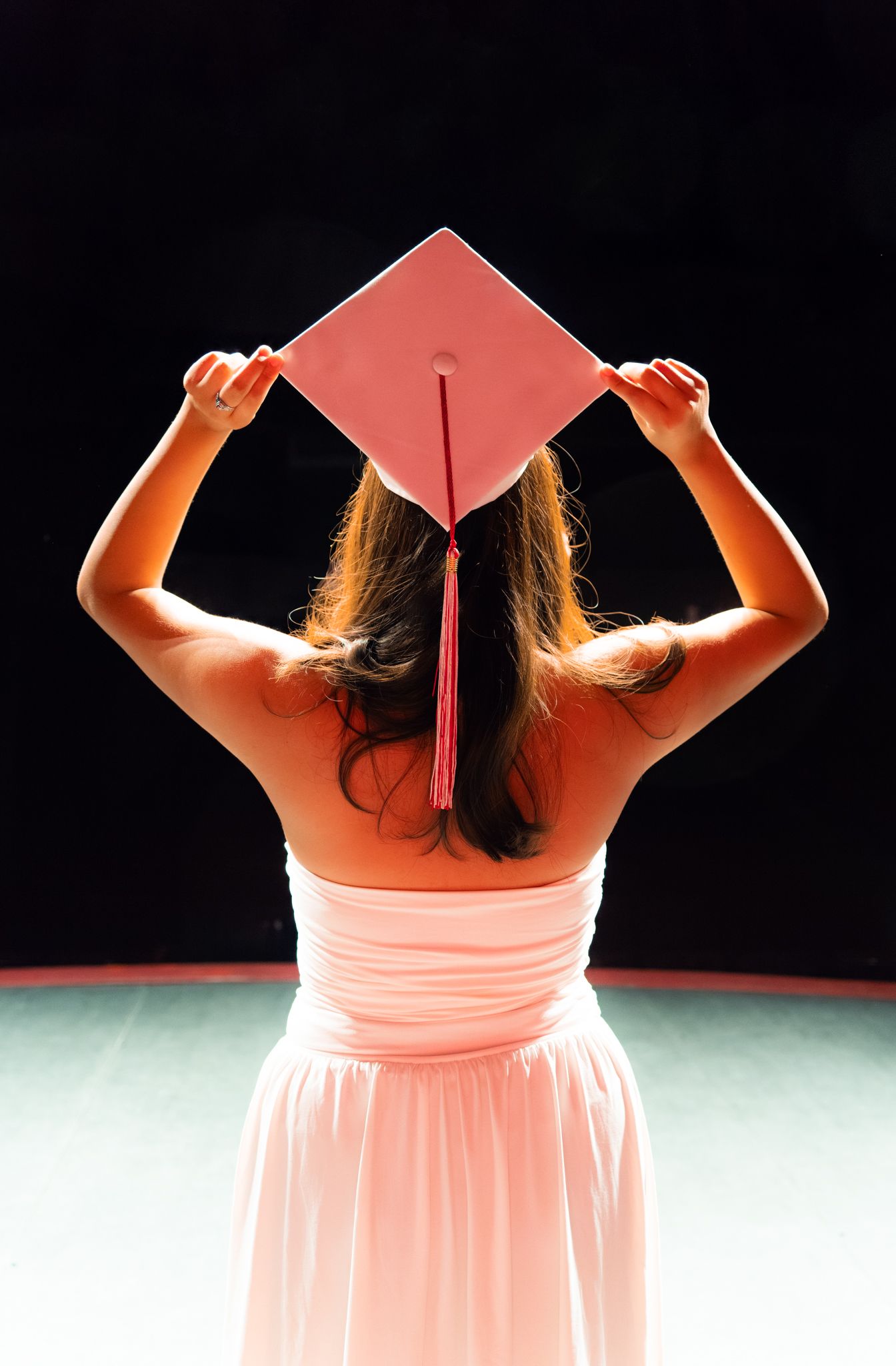 Young woman in pink dress holds red graduation cap above her head against a black background.