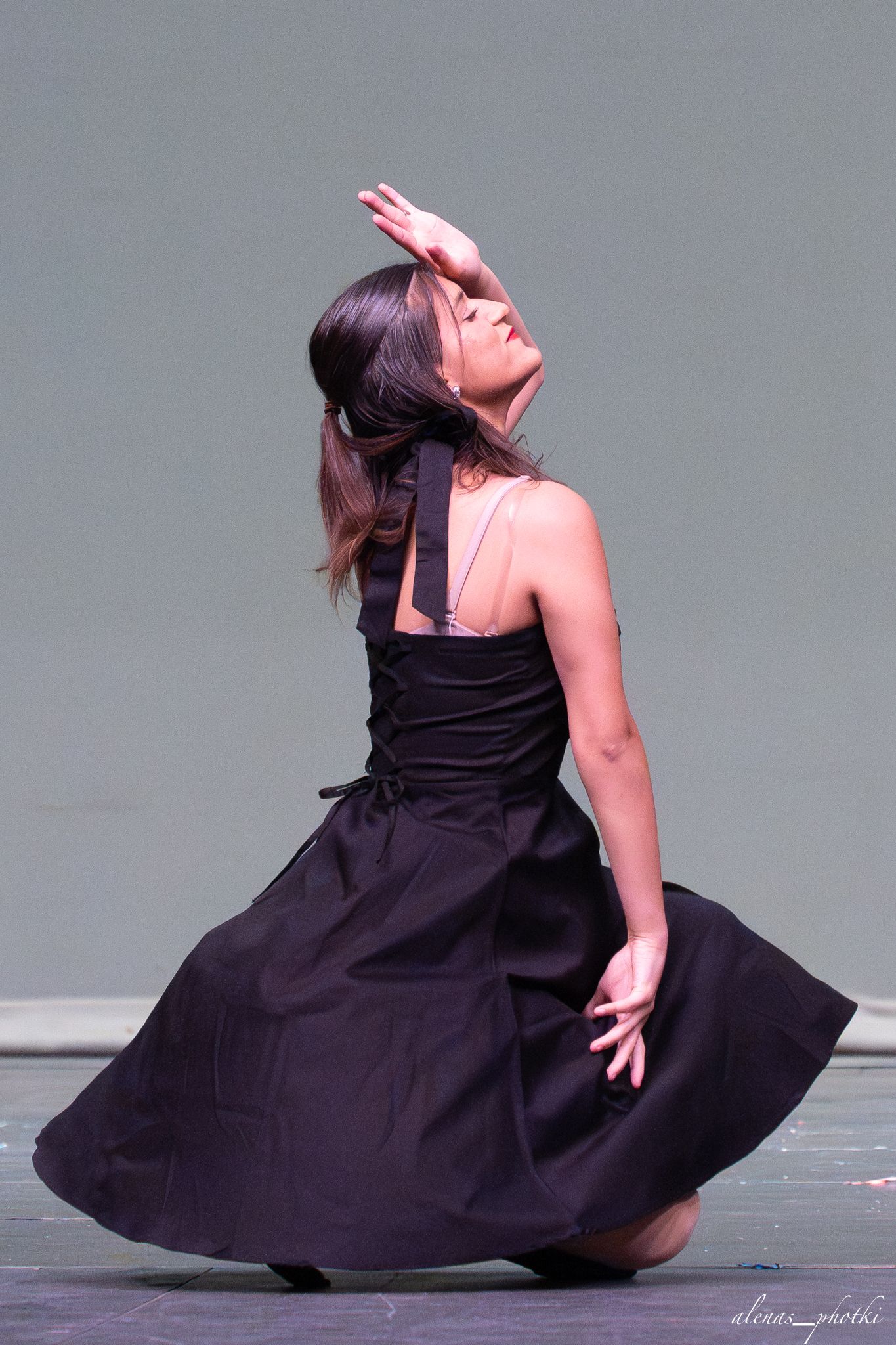 A female dancer in a black dress performs an expressive pose on stage, tilting her head back with one hand raised.