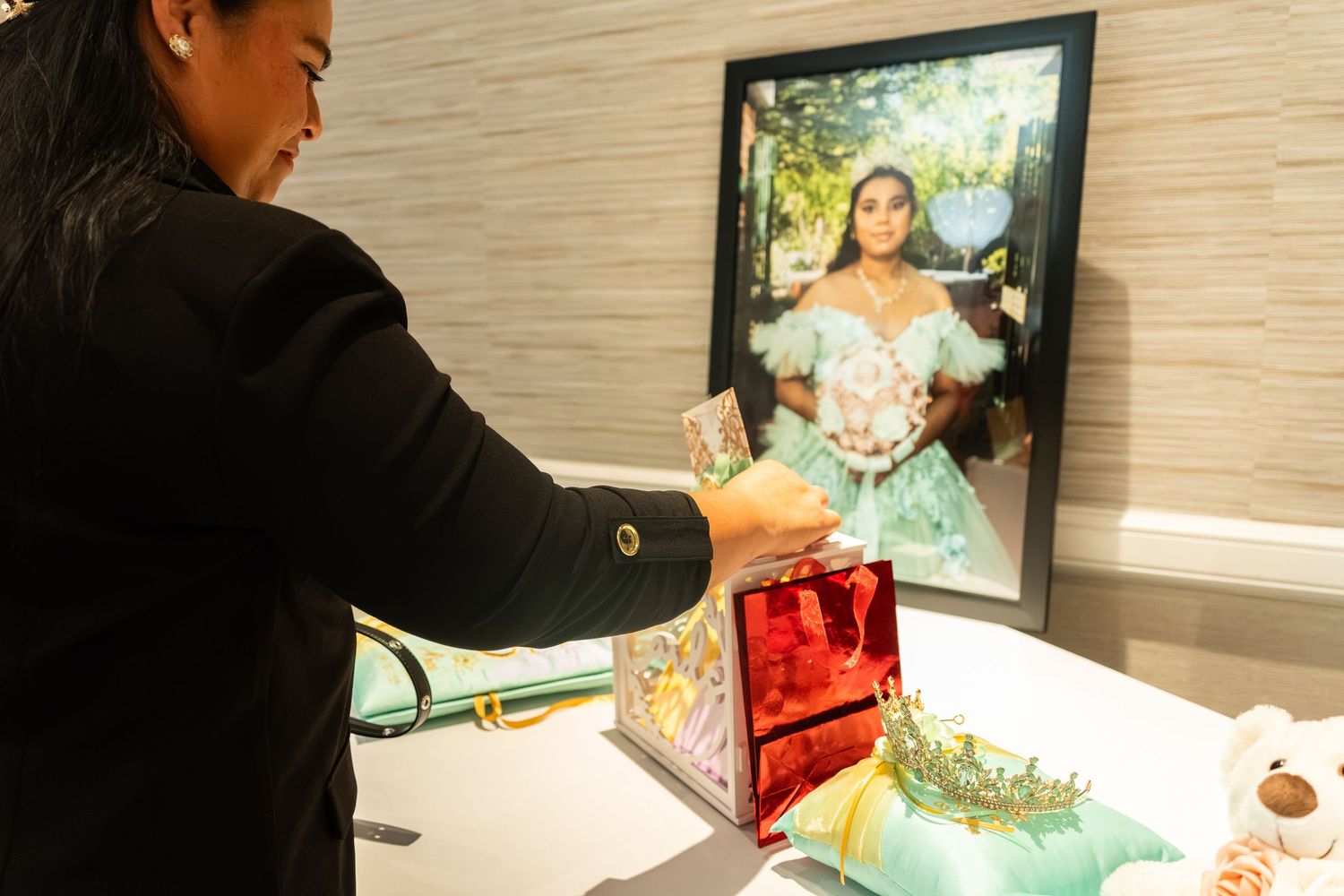 Woman arranges gifts on table beside framed quinceañera portrait in elegant mint green gown.