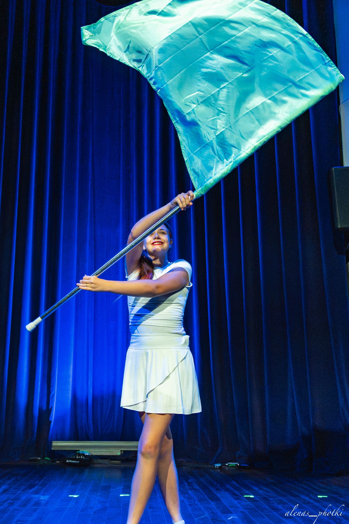 Performer in white dress spins turquoise fabric flags in sequence against blue stage curtain backdrop.