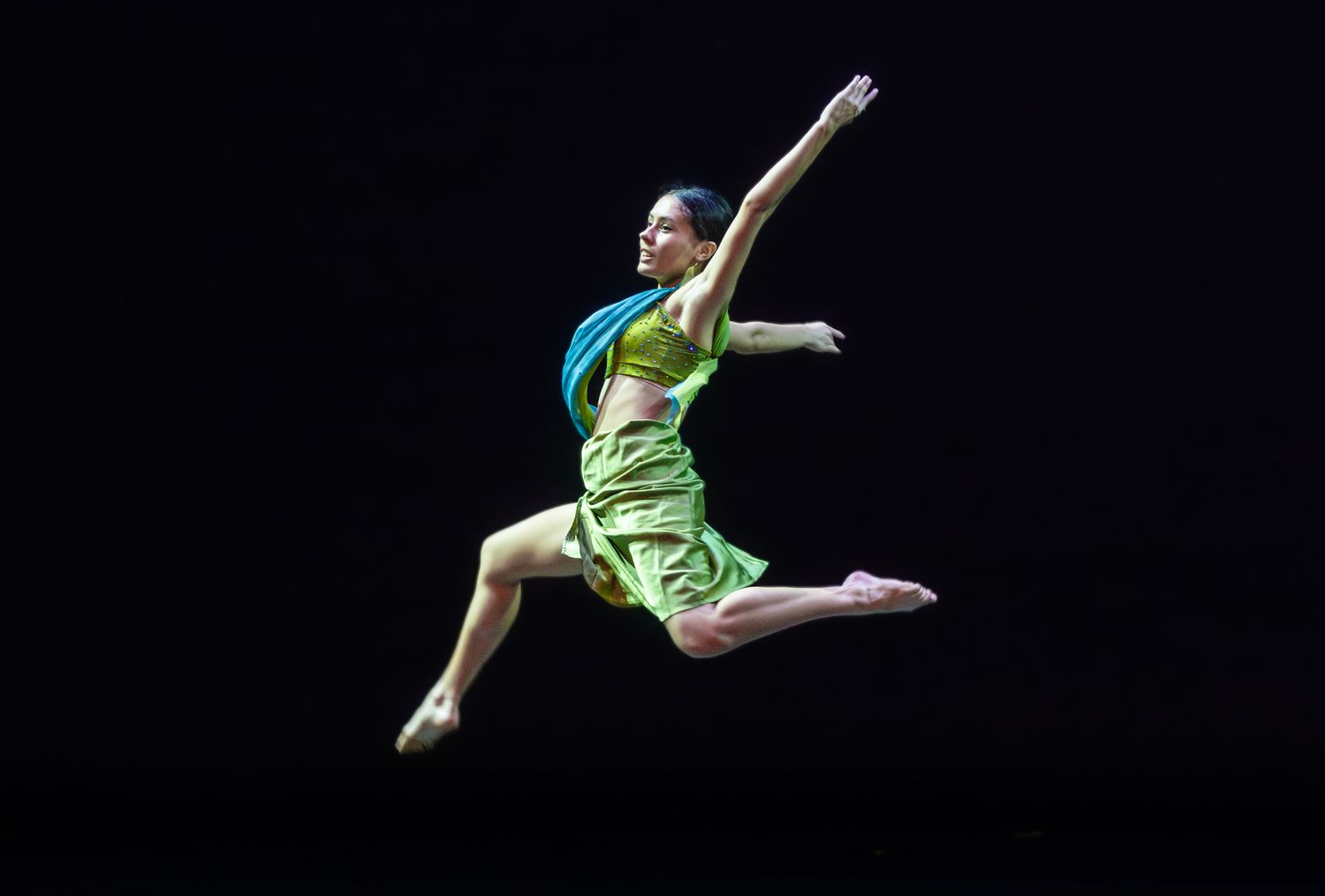 A dancer in a green dress leaps gracefully against a black background.