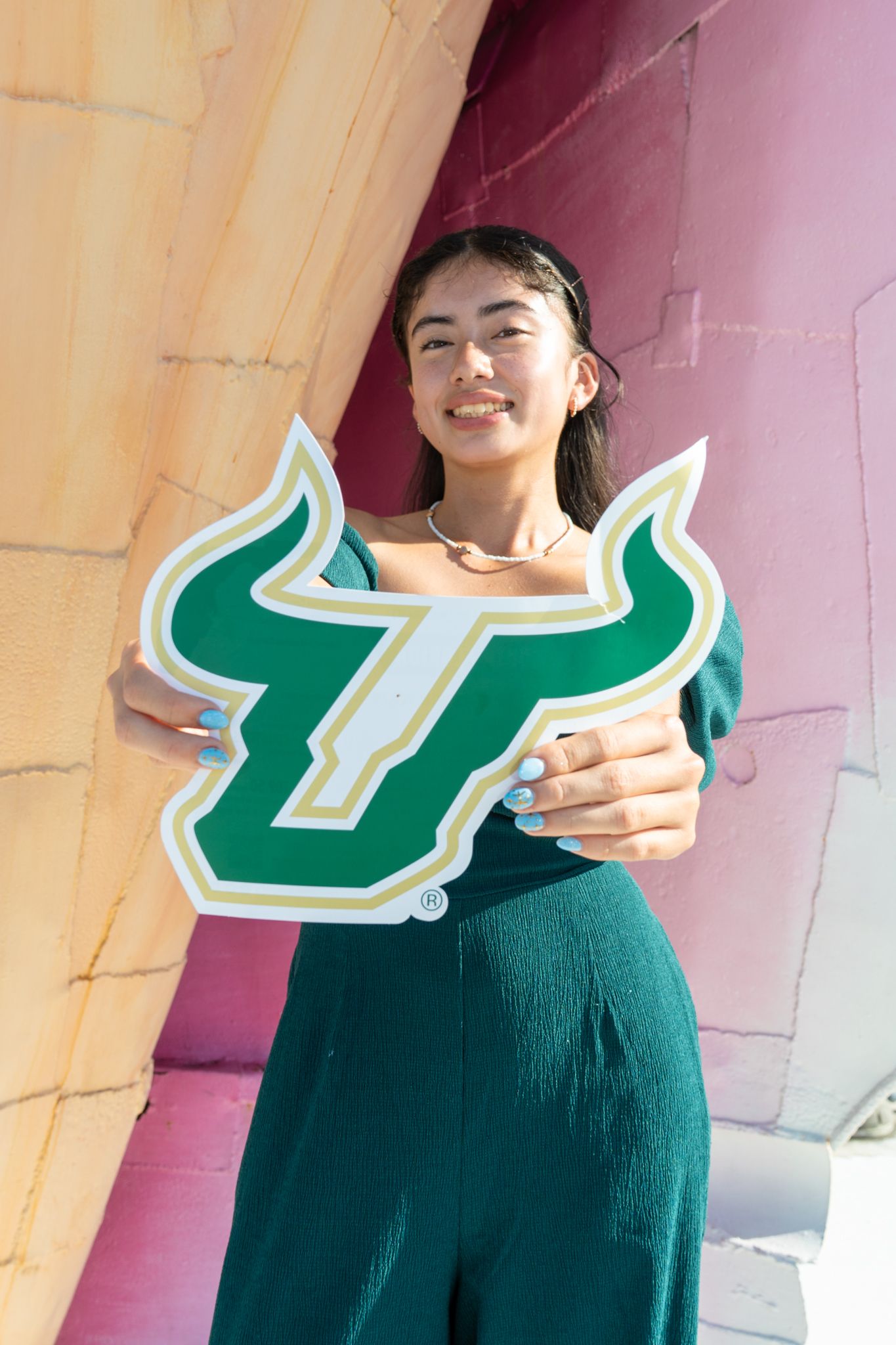 Smiling young woman holding a University of South Florida Bulls logo sign, wearing a green outfit outdoors.