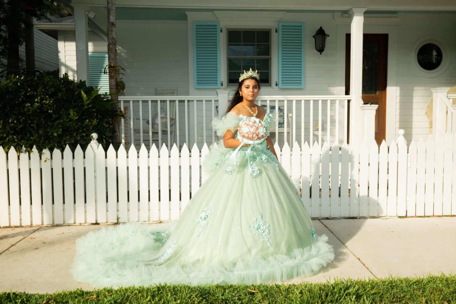 Young woman in mint green quinceañera ball gown with tiara holds cake outside white picket fence home.