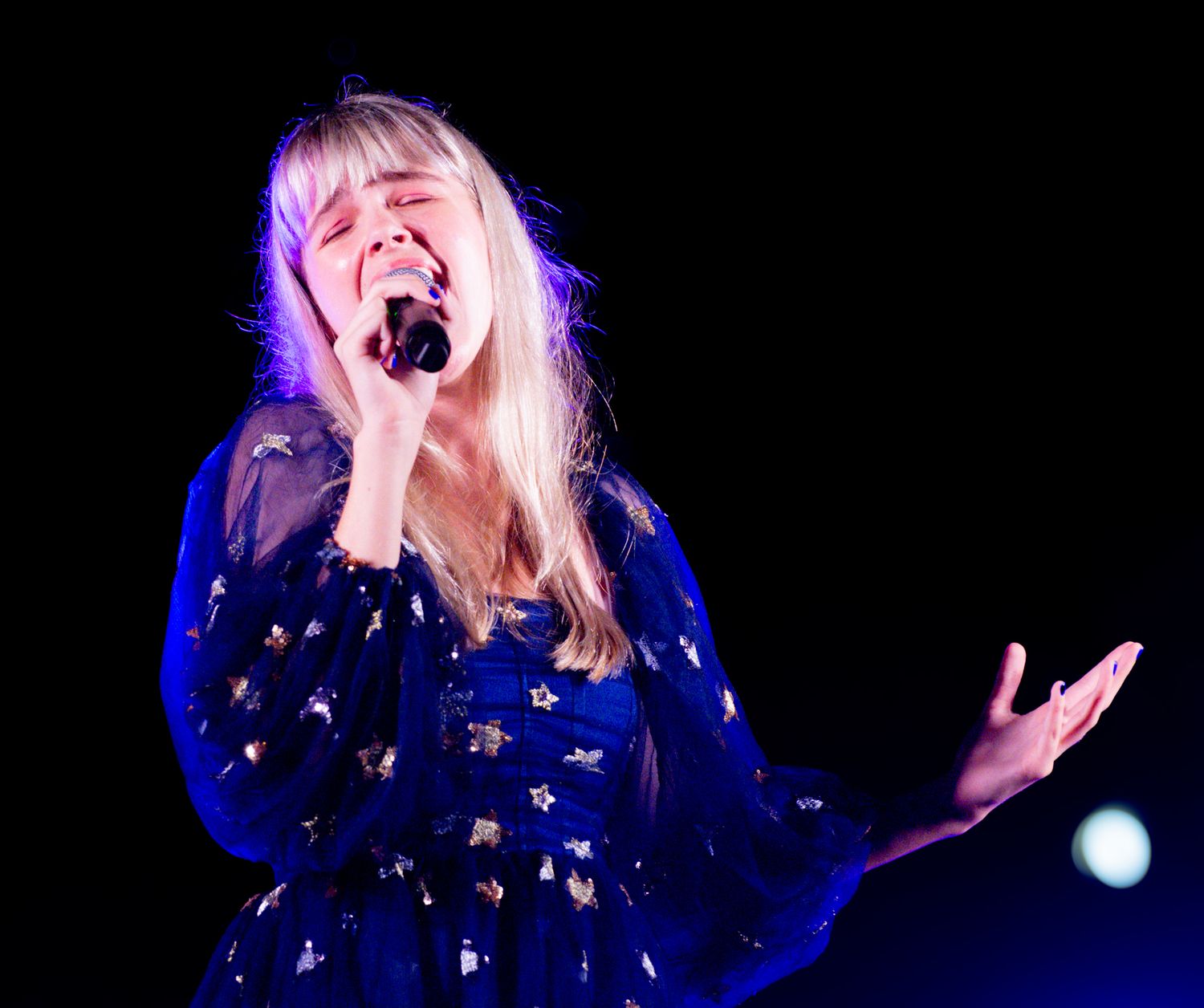 Singer in blue dress performing passionately against dark background with dramatic lighting.