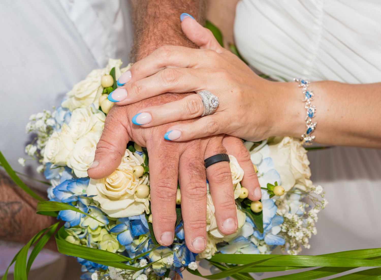 Close-up of hands with wedding rings resting on a bouquet of white and blue flowers.