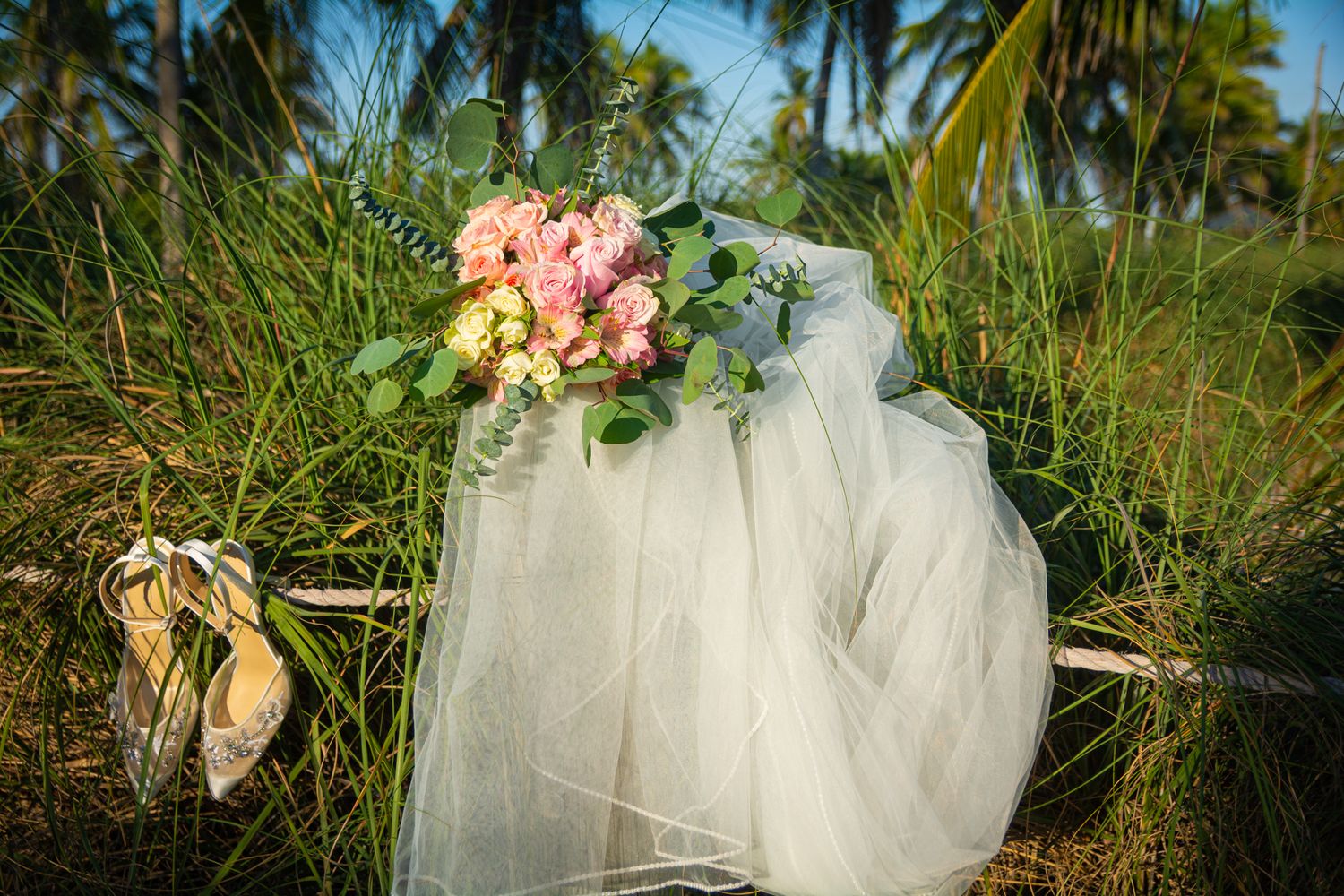 A wedding veil and bouquet with pink flowers resting in beach grass with palm trees.