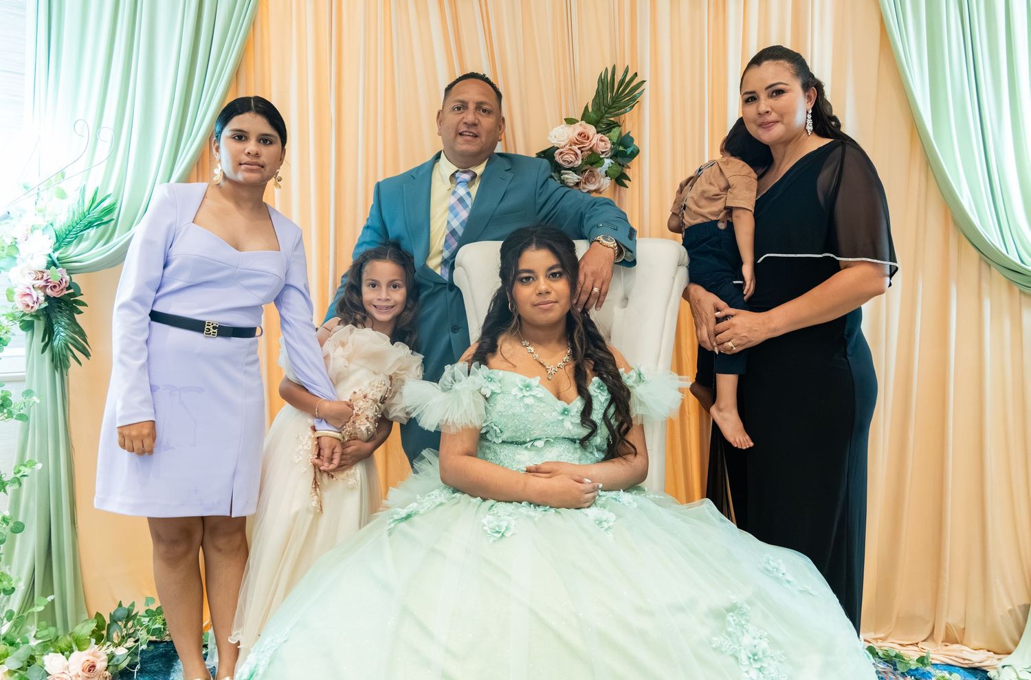 Quinceañera girl in mint ball gown poses with family including man, women, and baby at decorated backdrop.