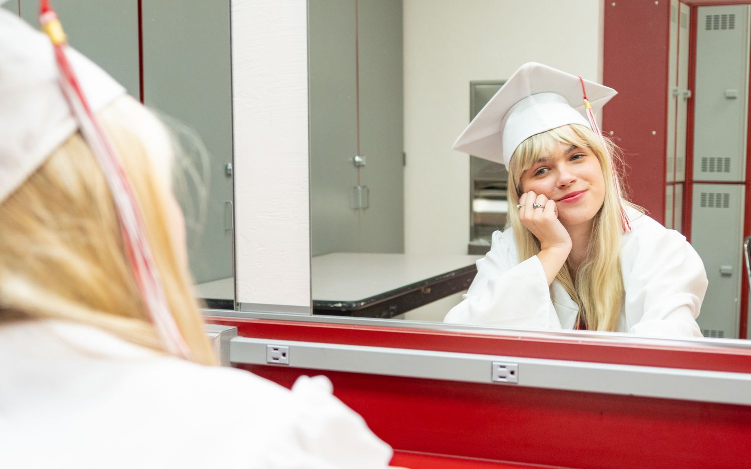 Blonde woman in white graduation cap and gown smiling at her reflection in a mirror near red lockers.