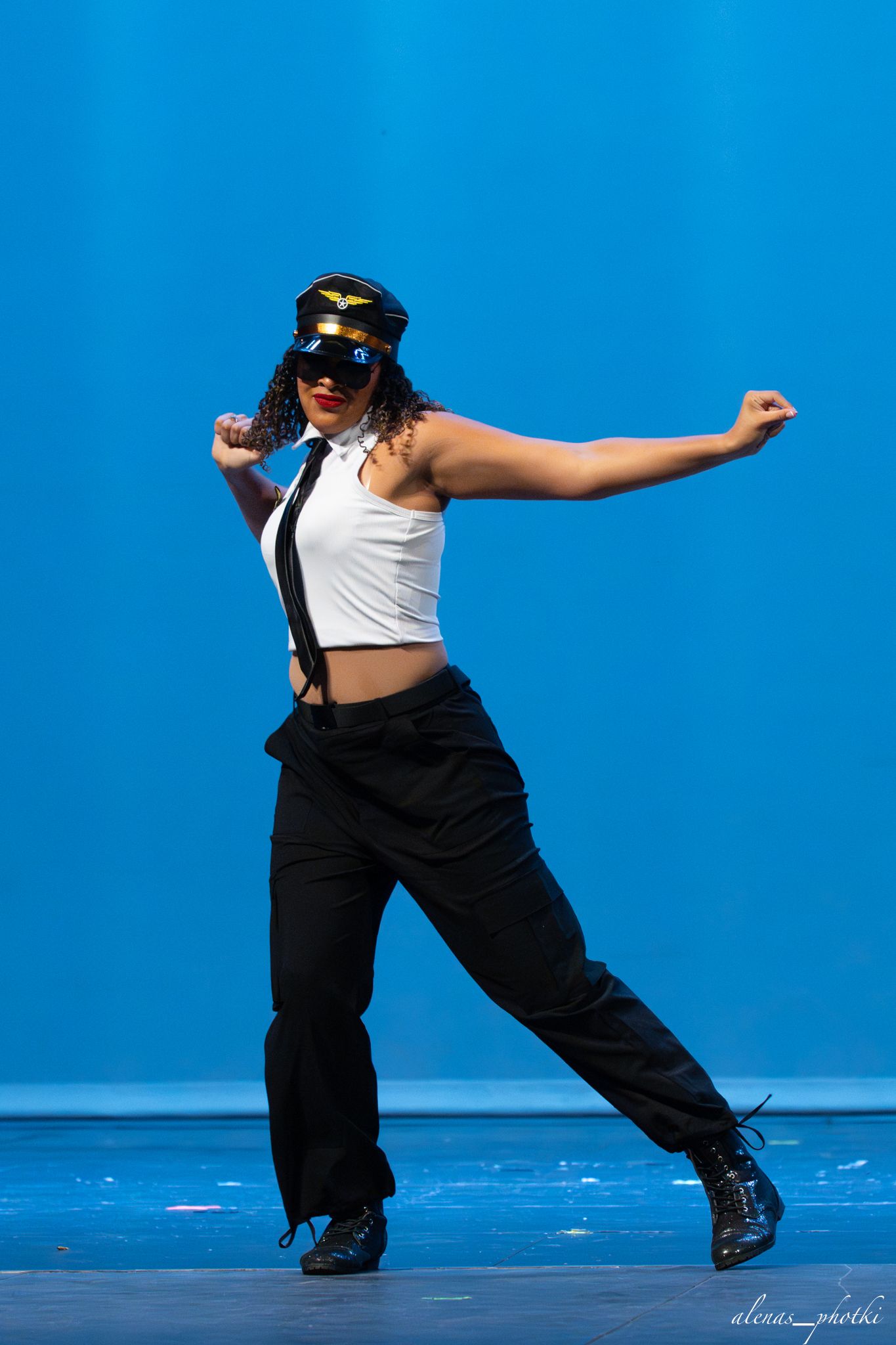 Dancer in pilot cap, white crop top, and black pants performs dynamic pose on blue stage.