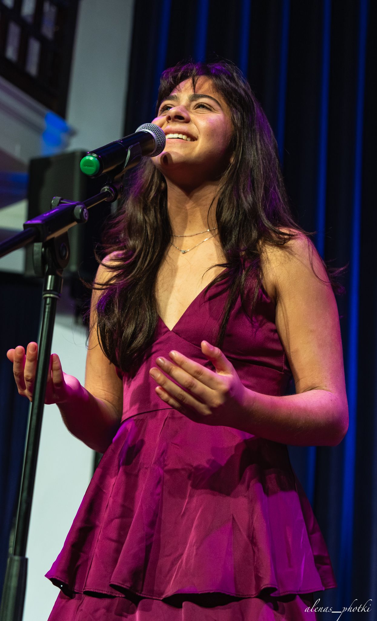 A singer in a flowing magenta dress performs emotively on stage with dramatic lighting and curtains.