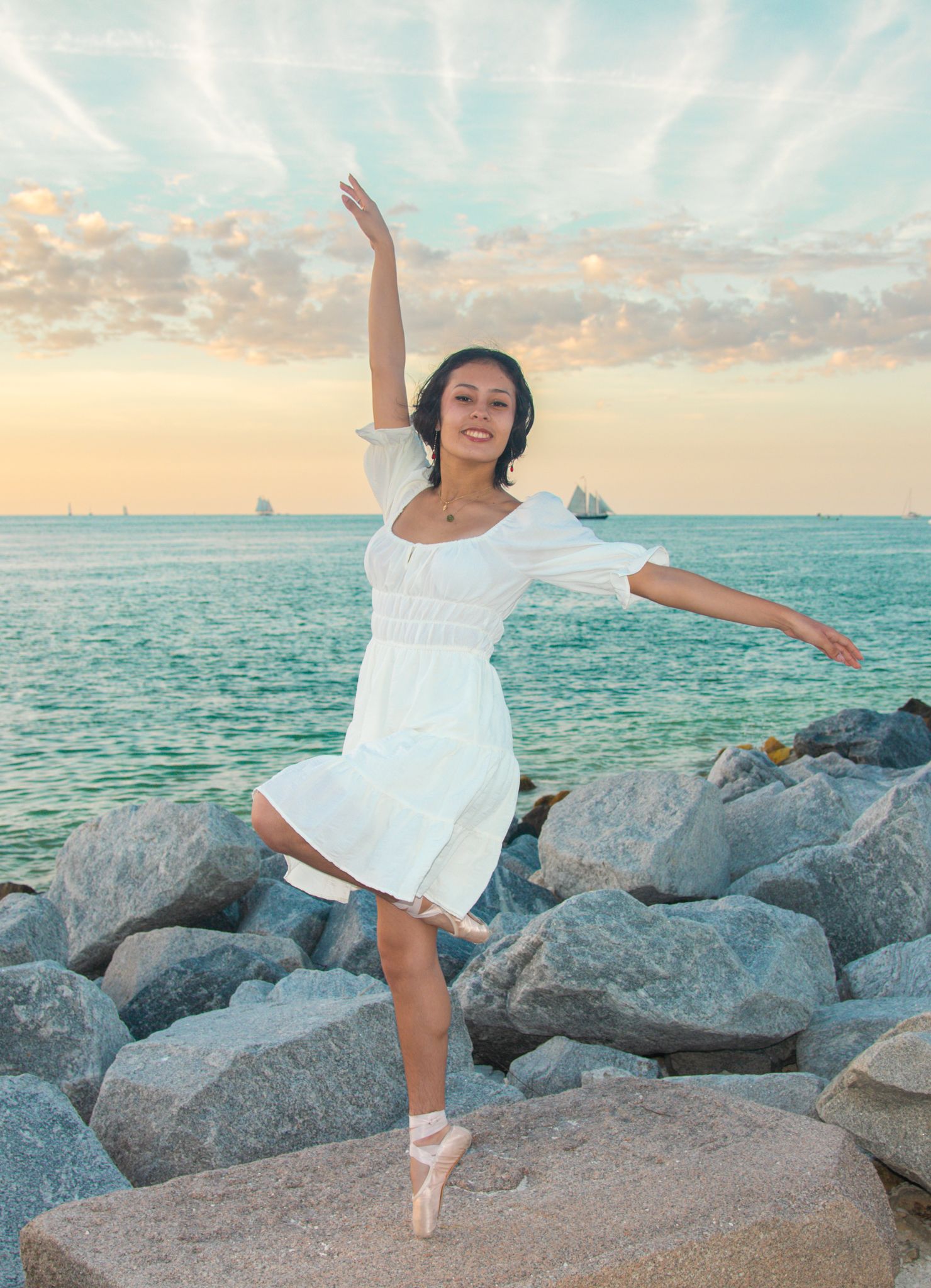 Ballet dancer in white dress poses on pointe among coastal rocks at dusk.