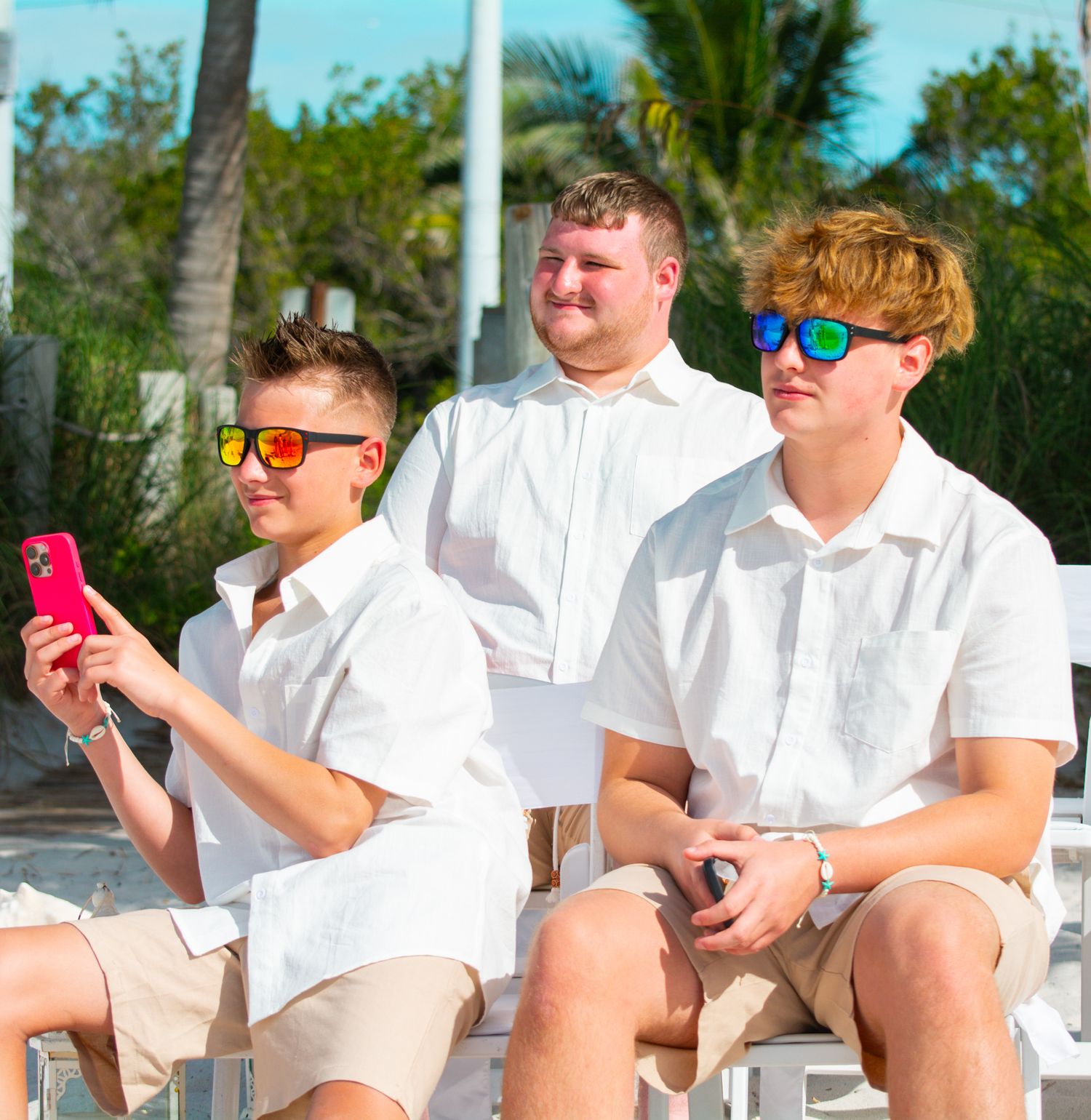 Three groomsmen in white shirts and khaki shorts sit casually on beach with sunglasses.