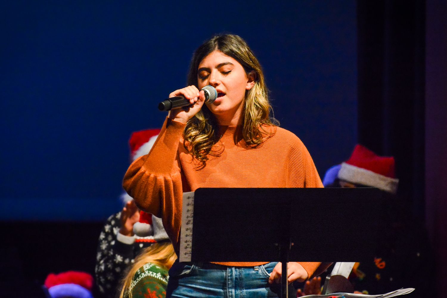 Performer in orange sweater sings at music stand during concert.
