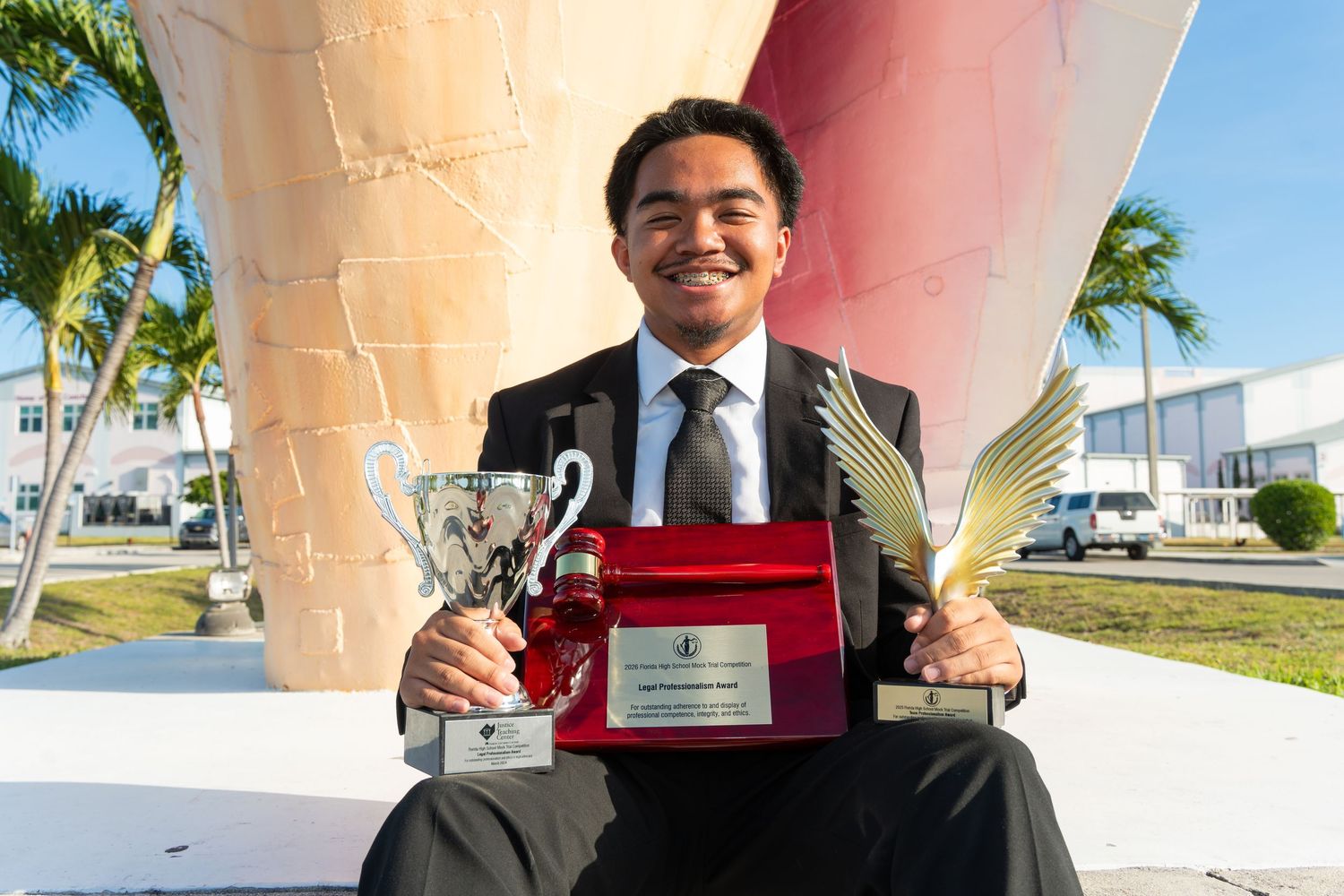 Young man in suit proudly holding trophy, award plaque, and golden palm statue outdoors near palm trees.