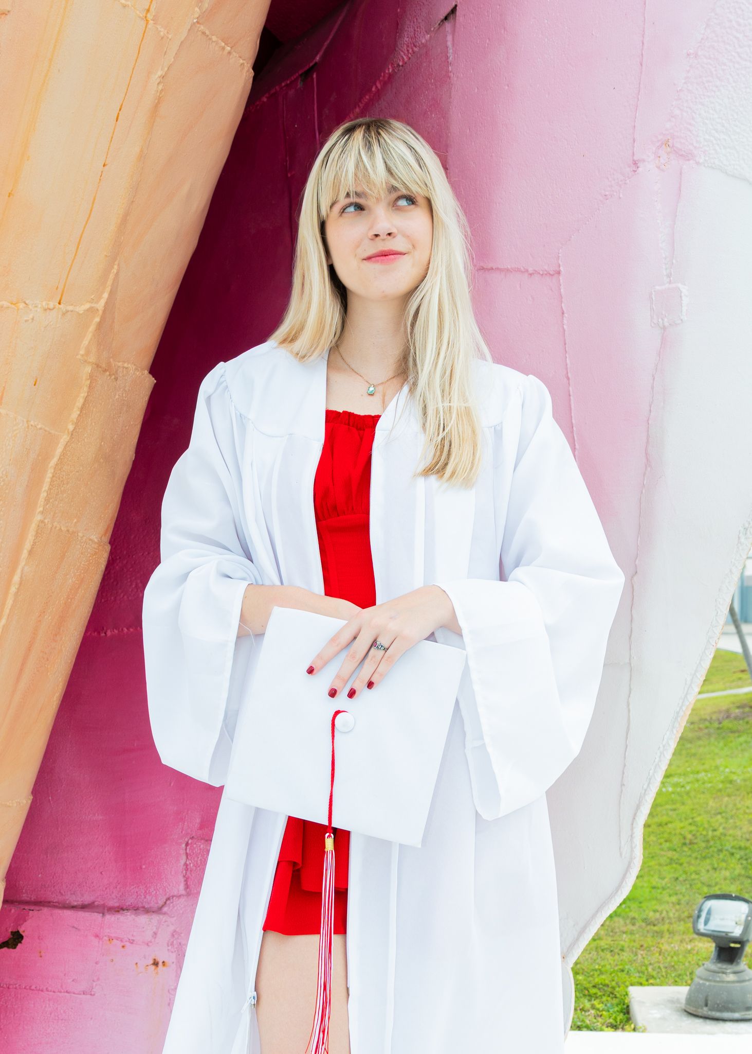 Young blonde woman in white graduation gown holding cap with red tassel, wearing red dress outdoors.