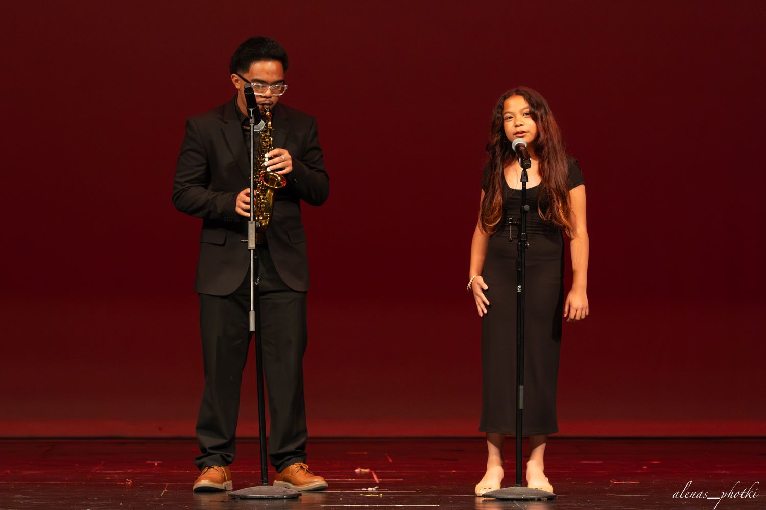 Two performers on stage, a man playing saxophone and a woman singing into microphones against a red backdrop.