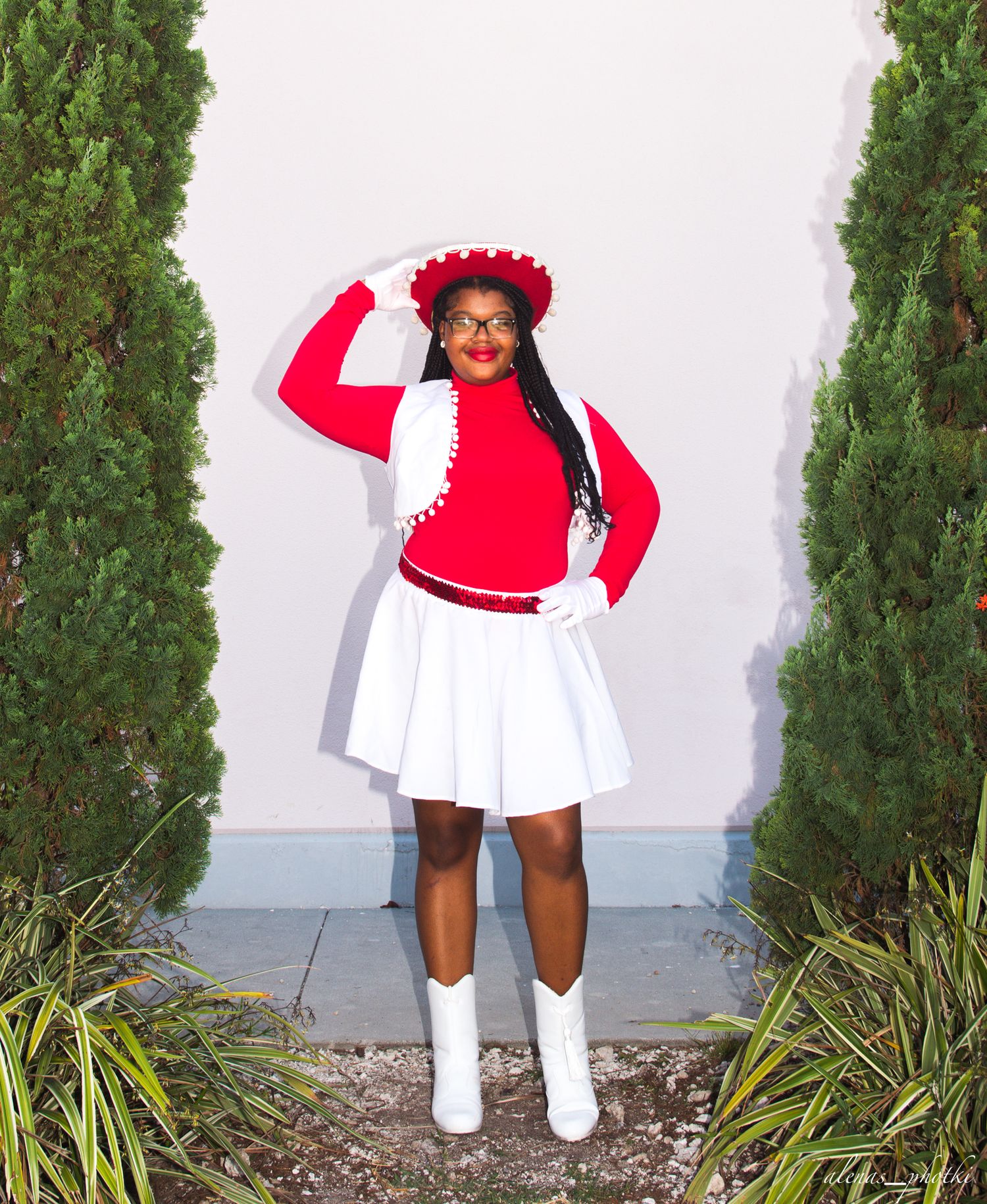 Cheerleader in red and white uniform with decorative hat poses between green shrubs.