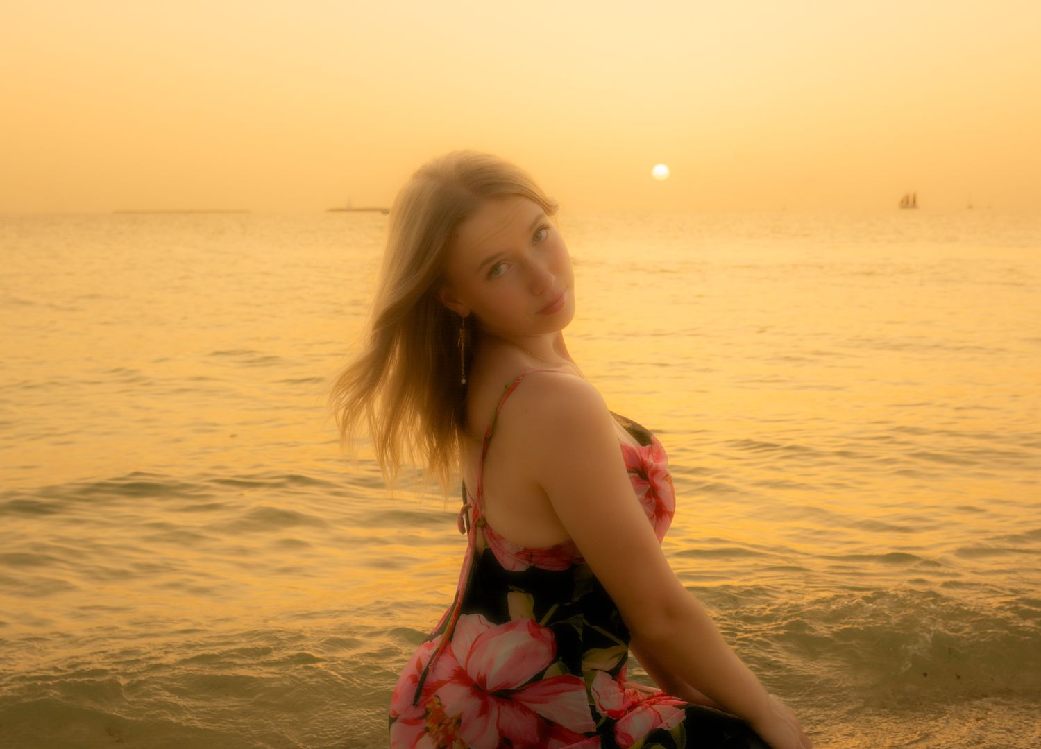 Woman in floral dress poses at golden hour by the ocean.