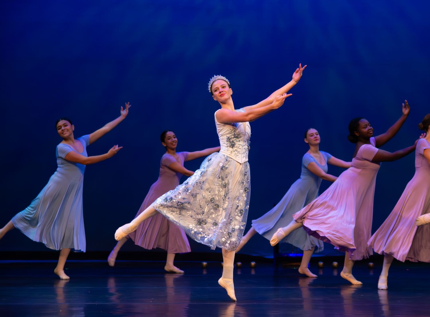 Ballet dancers in flowing dresses perform on stage against a deep blue backdrop.