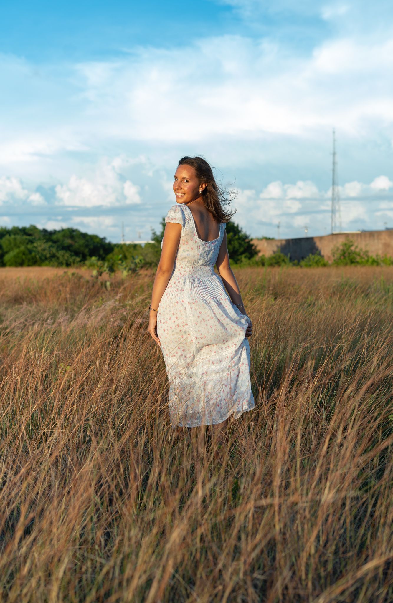 A flowing white dress catches the wind as someone twirls through tall golden grass under a blue summer sky.