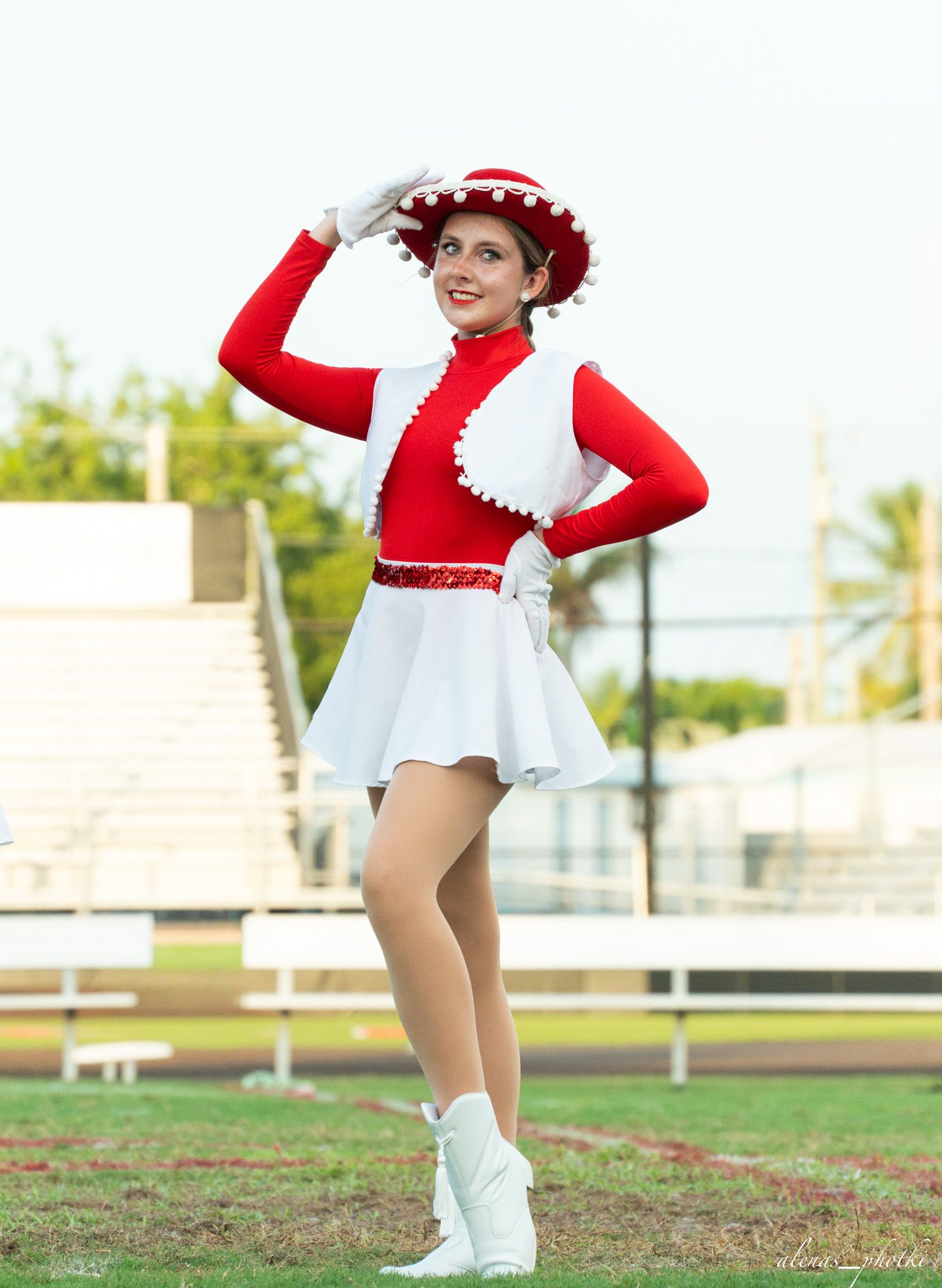 A smiling majorette in a red and white uniform with a decorative hat performs on a football field.