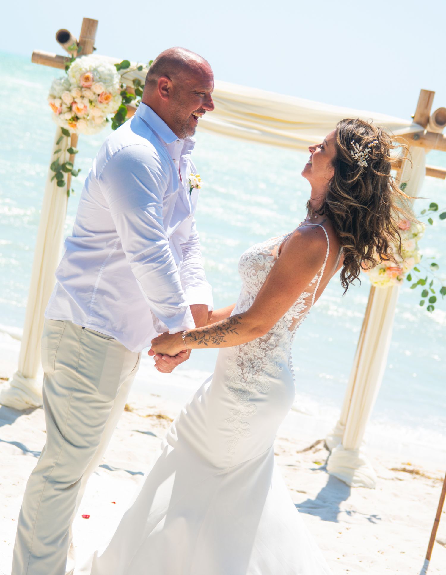 A couple shares a joyful moment during their seaside wedding ceremony under a white arch.