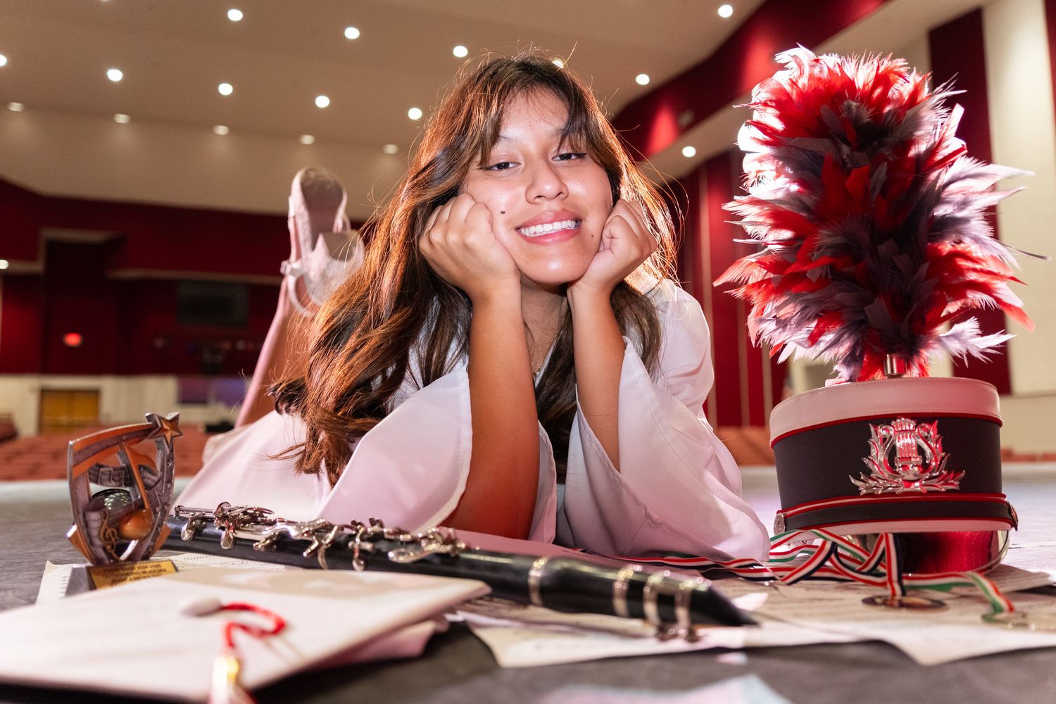 Smiling young woman poses with a marching band hat, clarinet, and sheet music in a red auditorium.
