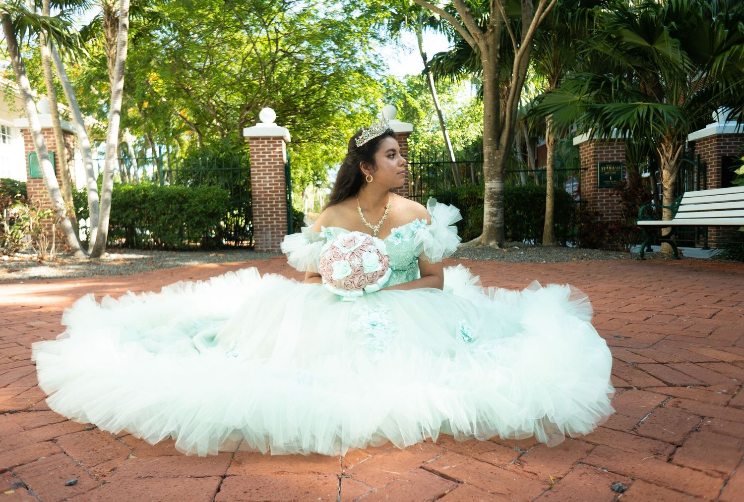 Young woman in a mint green quinceañera ball gown poses on brick courtyard surrounded by lush greenery.