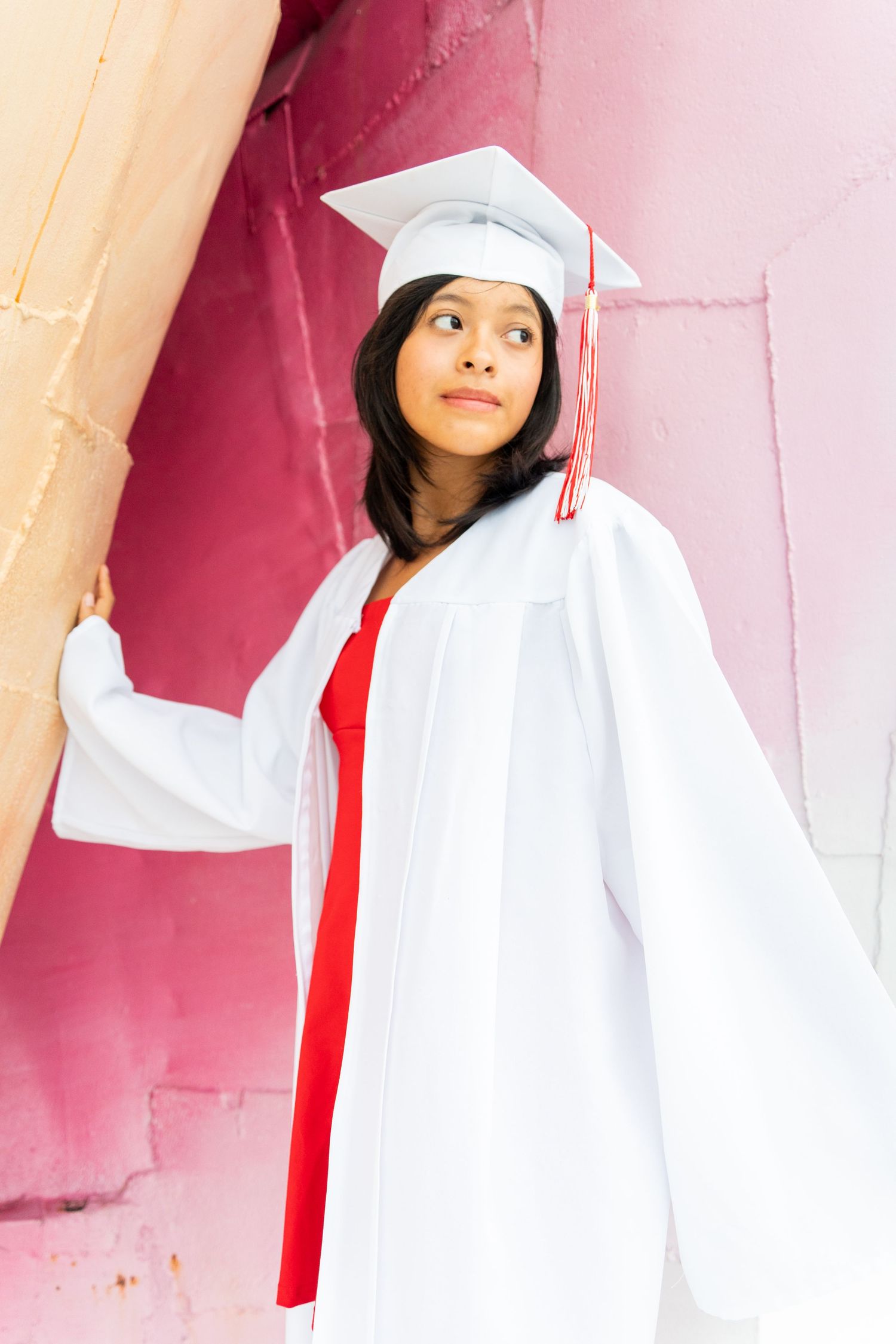 Young woman in white graduation cap and gown posing confidently against a pink wall.