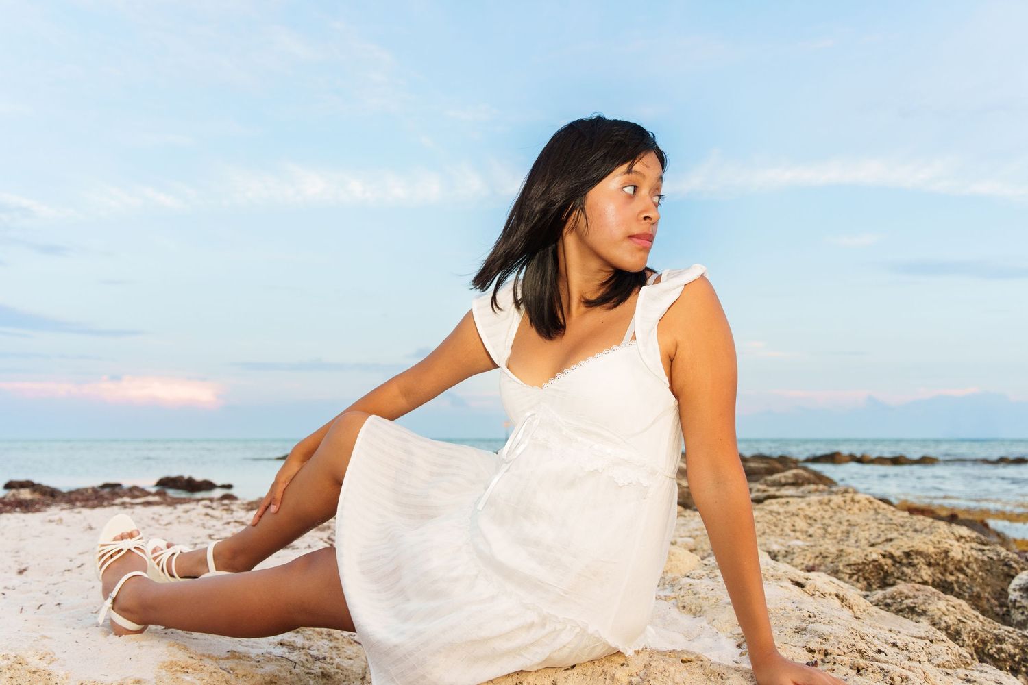 A young woman in a white dress sits on rocks at a beach, gazing sideways with the ocean in the background.