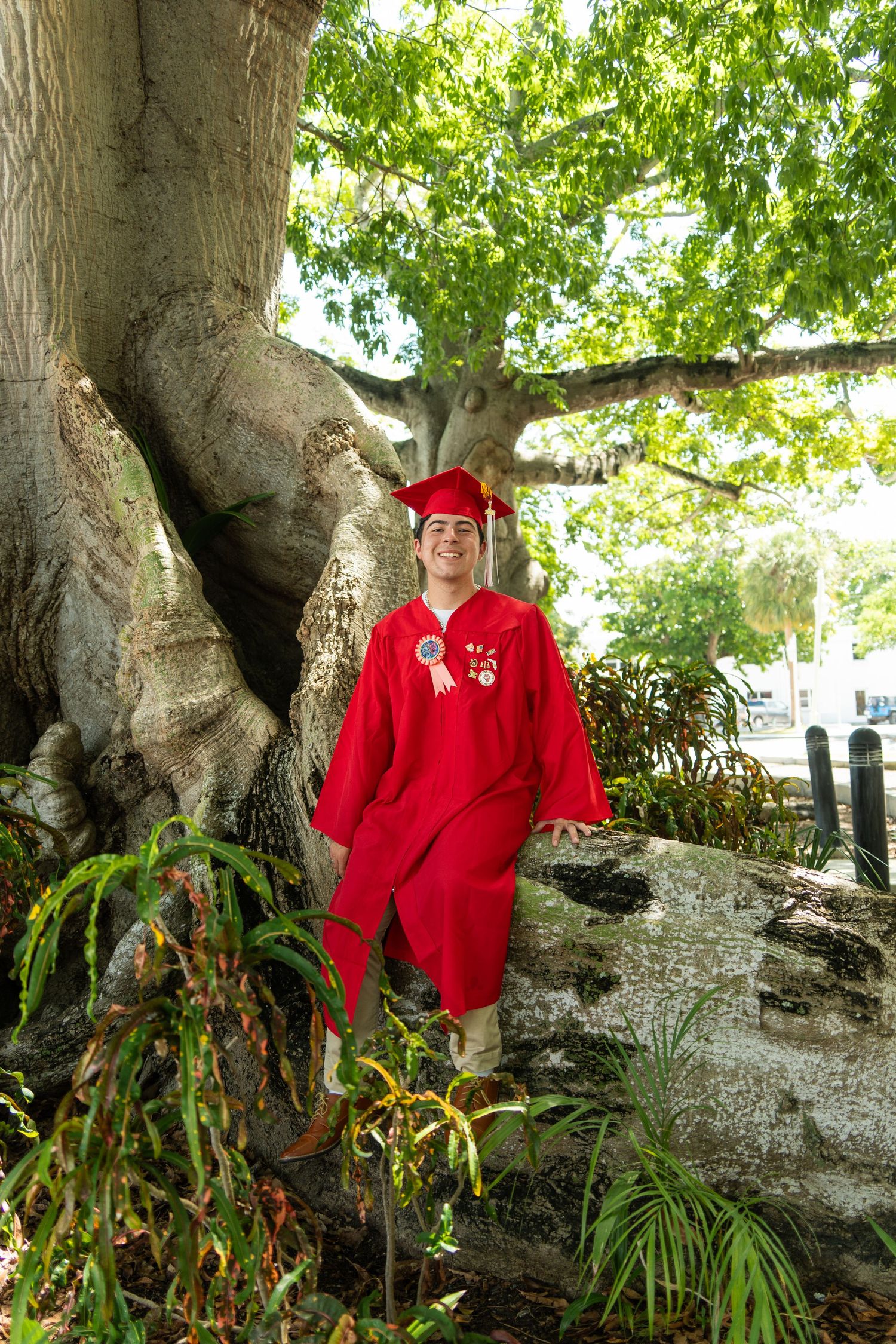 Graduate in red cap and gown smiling beside a large tree with exposed roots in a lush garden setting.