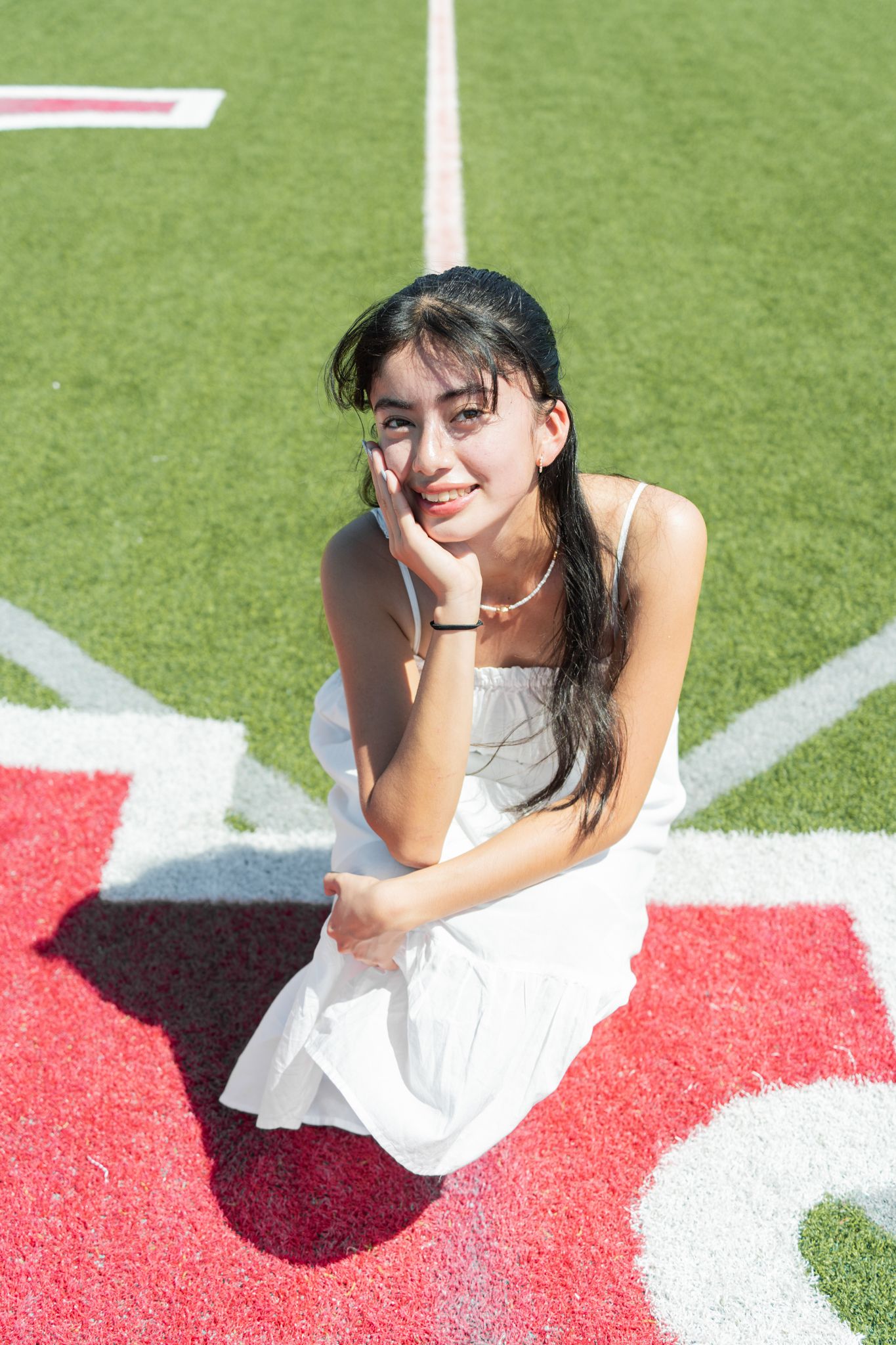 Young woman in white dress sitting on red and green turf football field, smiling with hand on cheek.