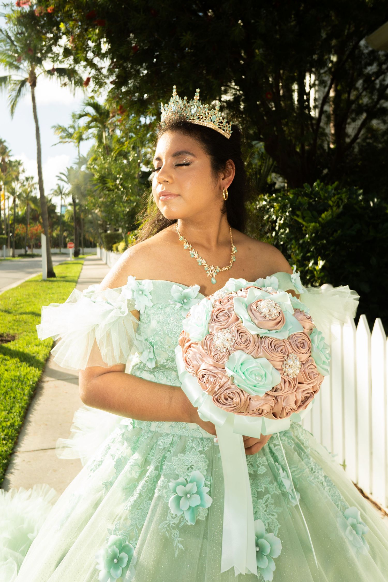 Teen in mint quinceañera gown and crown holds pink rose bouquet outdoors near white picket fence.