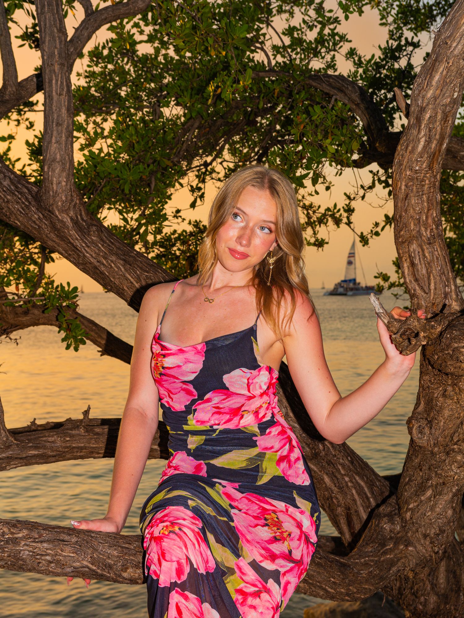 Woman in floral dress poses on tree branch against golden sunset sky.