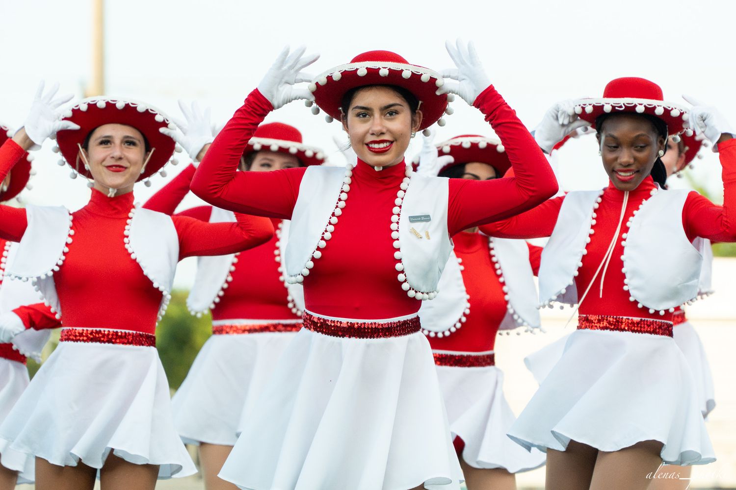 Smiling majorettes in red and white uniforms with matching hats perform a dance routine outdoors.