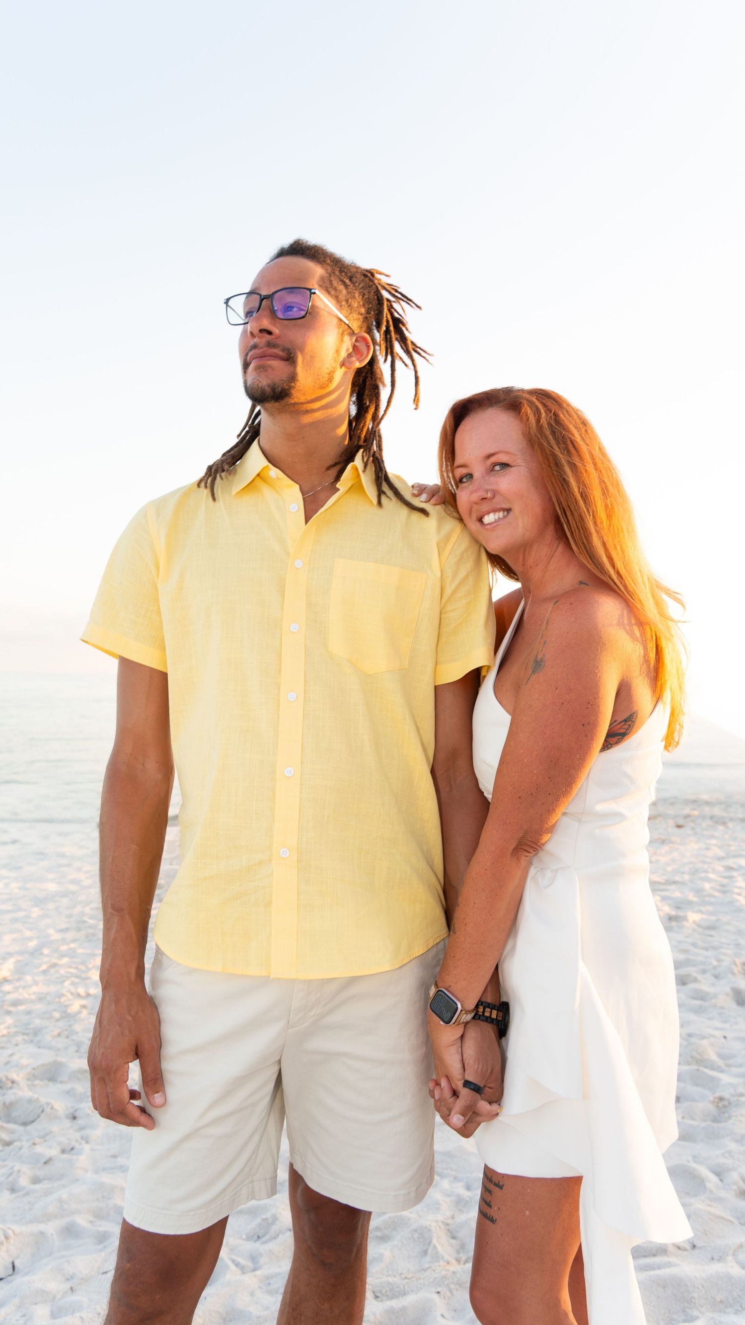 A couple holds hands on a sunny beach, man in yellow shirt with dreadlocks, woman in white dress with red hair.