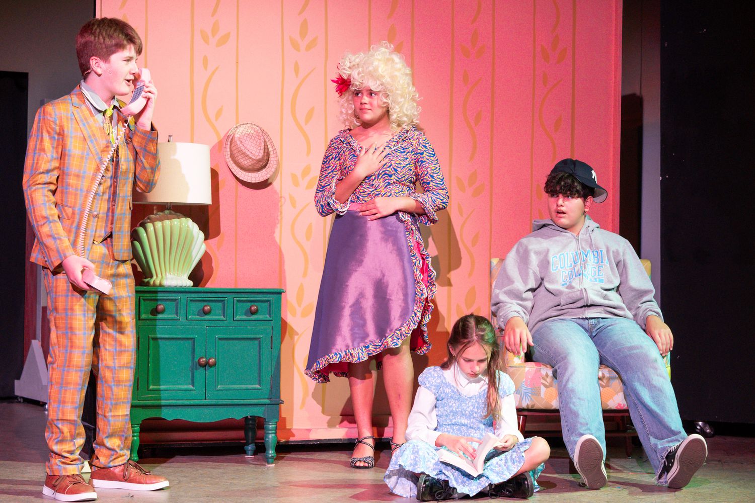 Theater stage performance with colorful costumes and props against pink backdrop.