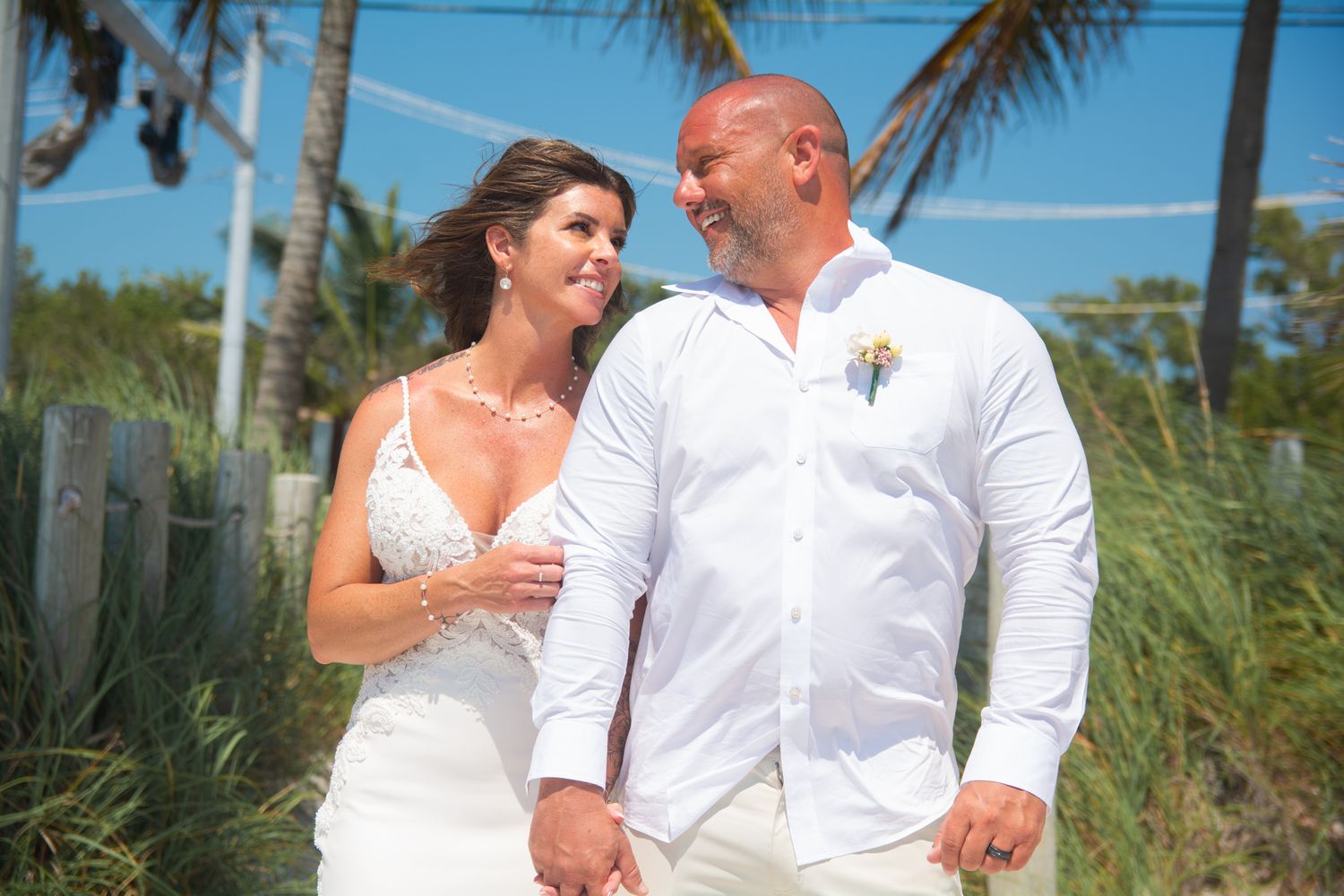 A couple in white wedding attire smiling together against a backdrop of palm trees.