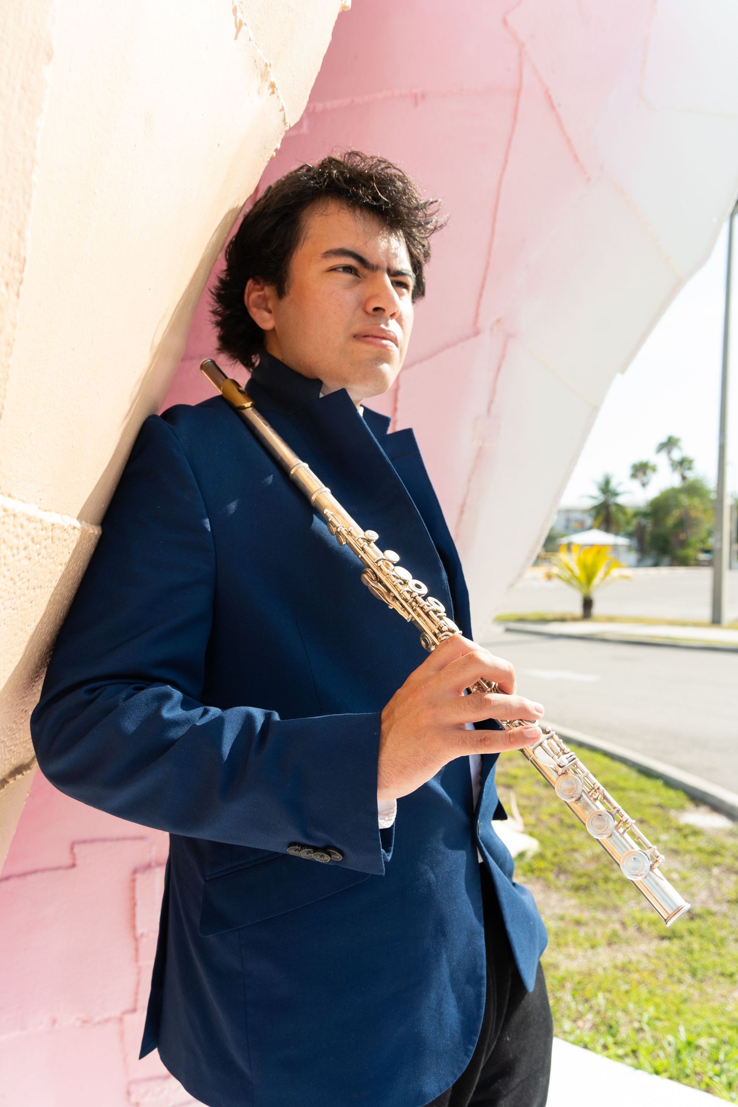 Young musician in blue blazer holding a flute outdoors near a pink wall on a sunny day.