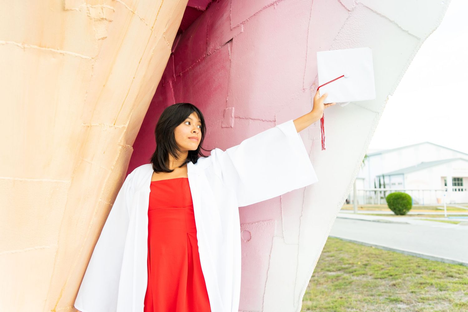 Young woman in white graduation gown and red dress poses outdoors near colorful wall holding diploma.