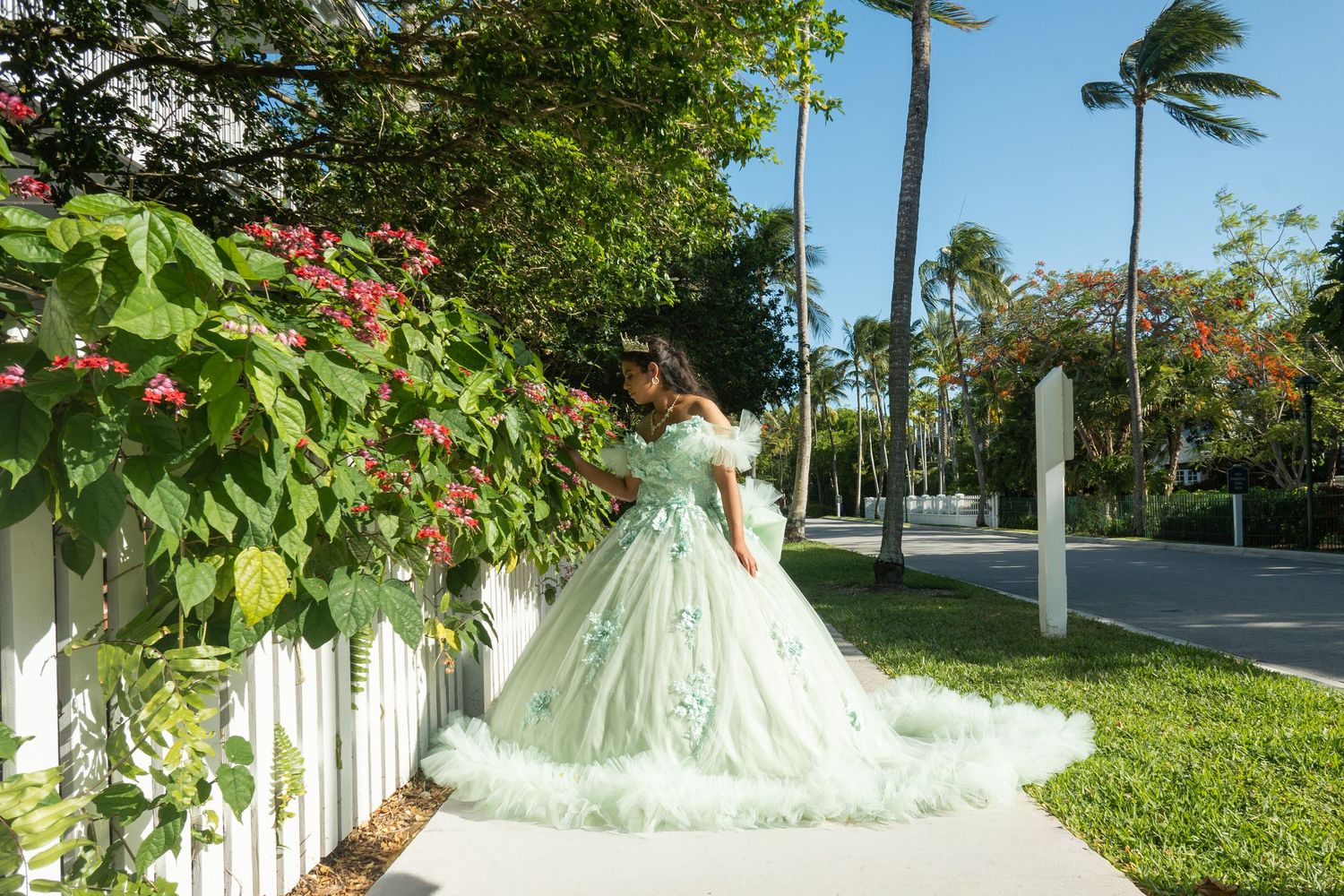 Young woman in mint green quinceañera ball gown poses by white fence with tropical flowers and palm trees.