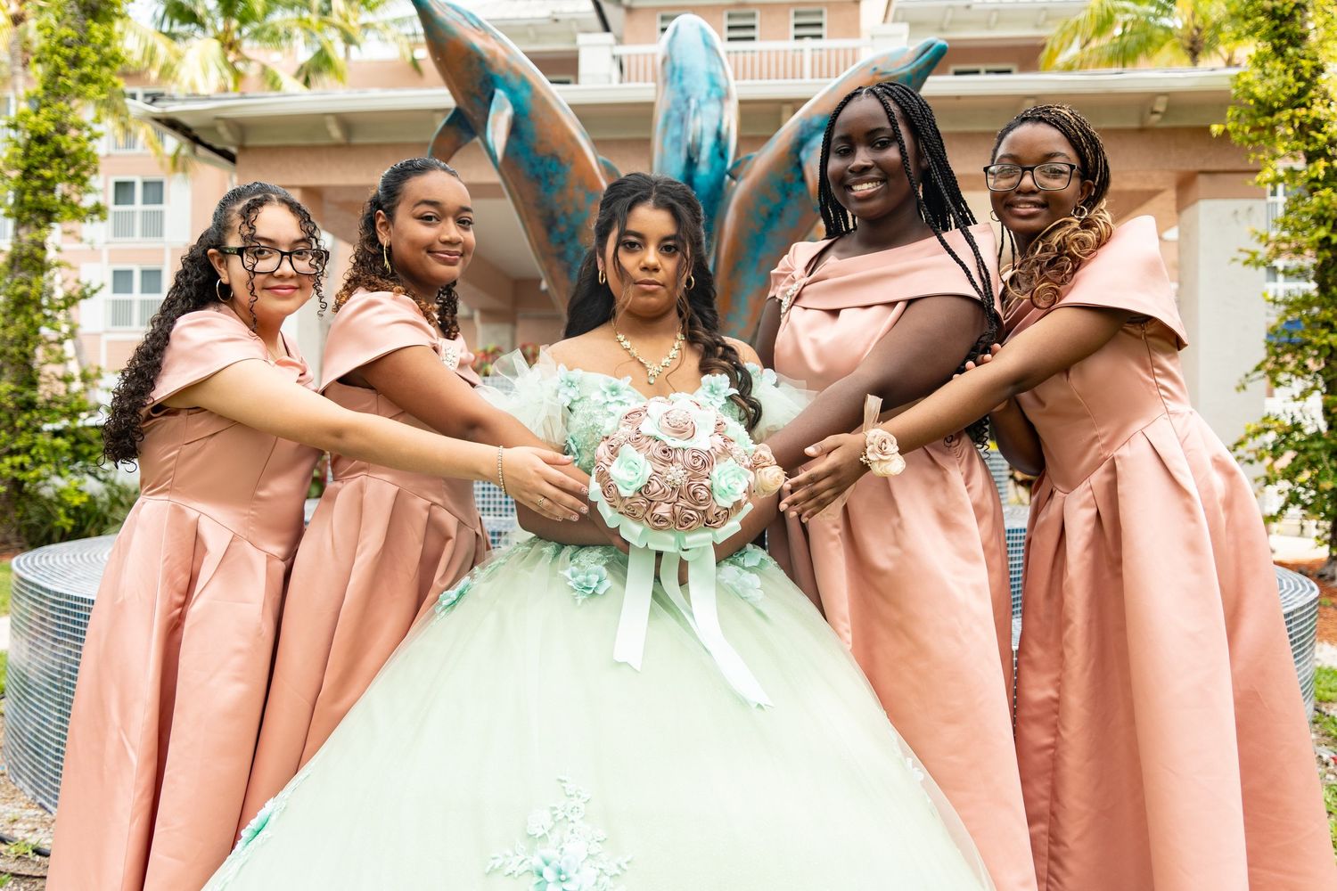 Five young women in pink dresses pose with a quinceañera girl in a mint green ball gown holding a bouquet.