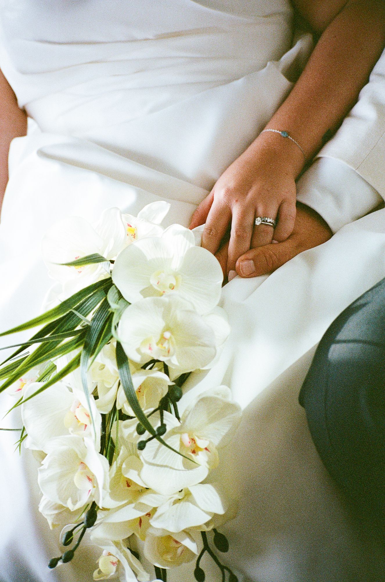 A beautiful white orchid bouquet rests against a soft white fabric background with a glimpse of an elegant wedding ring.
